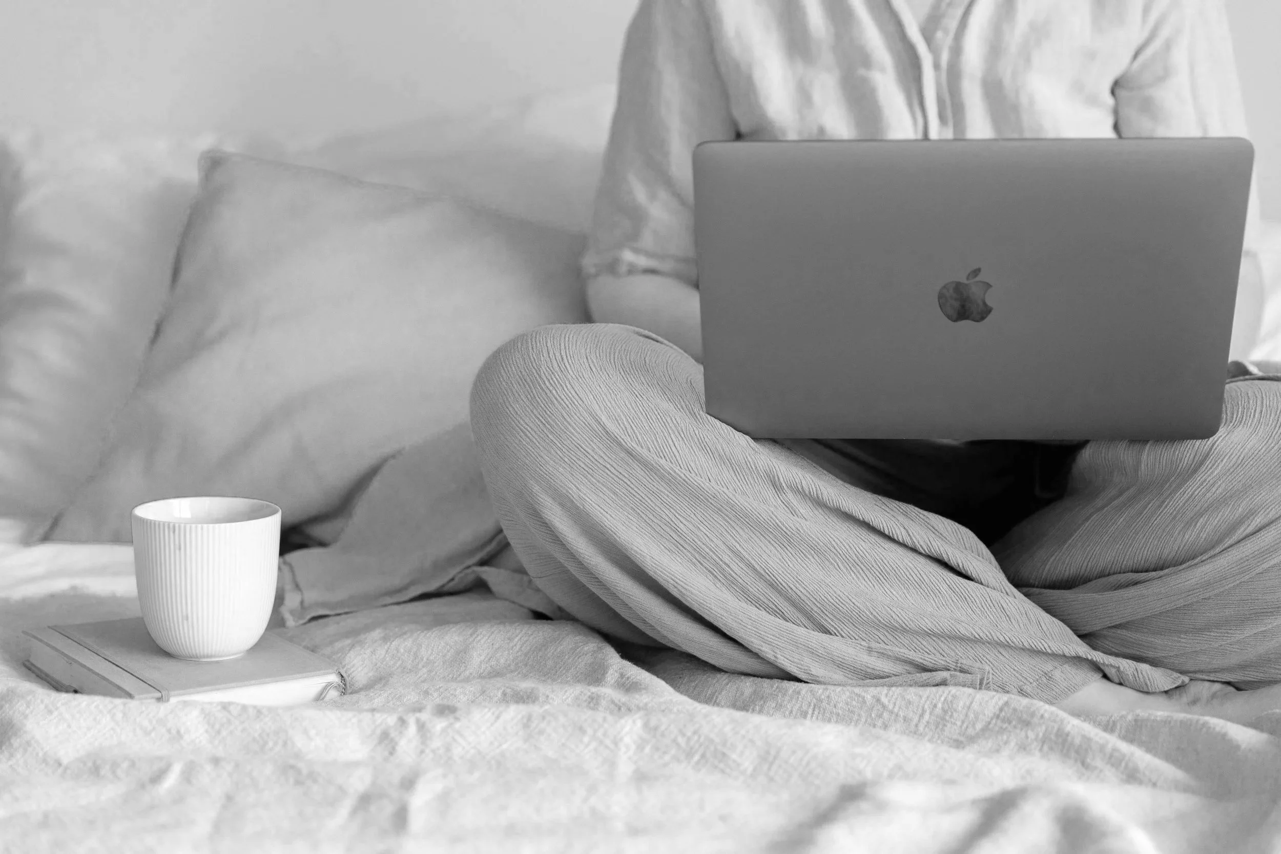A woman working on her laptop in bed on a web design project, sitting next to a book and a cup of tea.