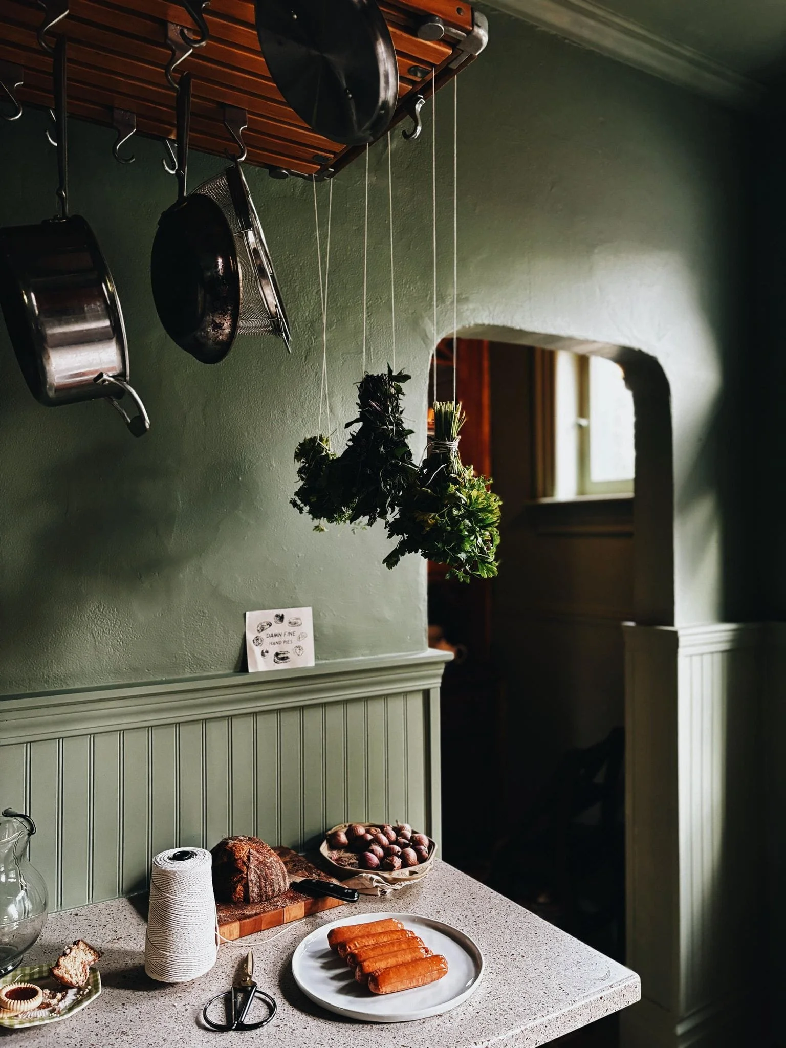 Dark green farm house kitchen with fresh herbs and pots hanging, and fresh bread and other ingredients on the counter.