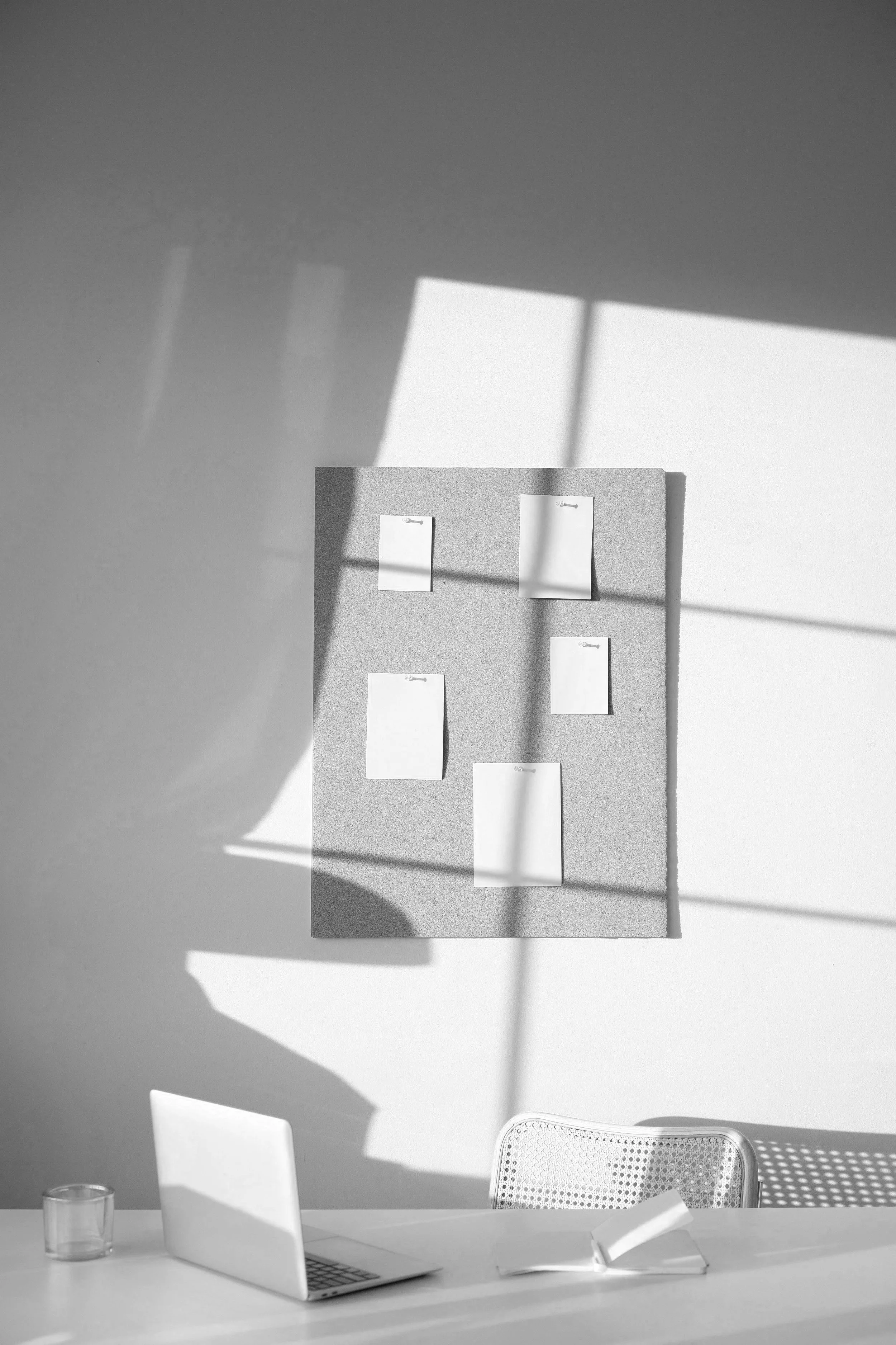 A minimalist, modern workspace showing a bulletin board on the wall with blank pages, an open laptop on the table with a coffee mug, and a wicker chair. There's a shadow being cast on the wall from morning light of a window and curtains.