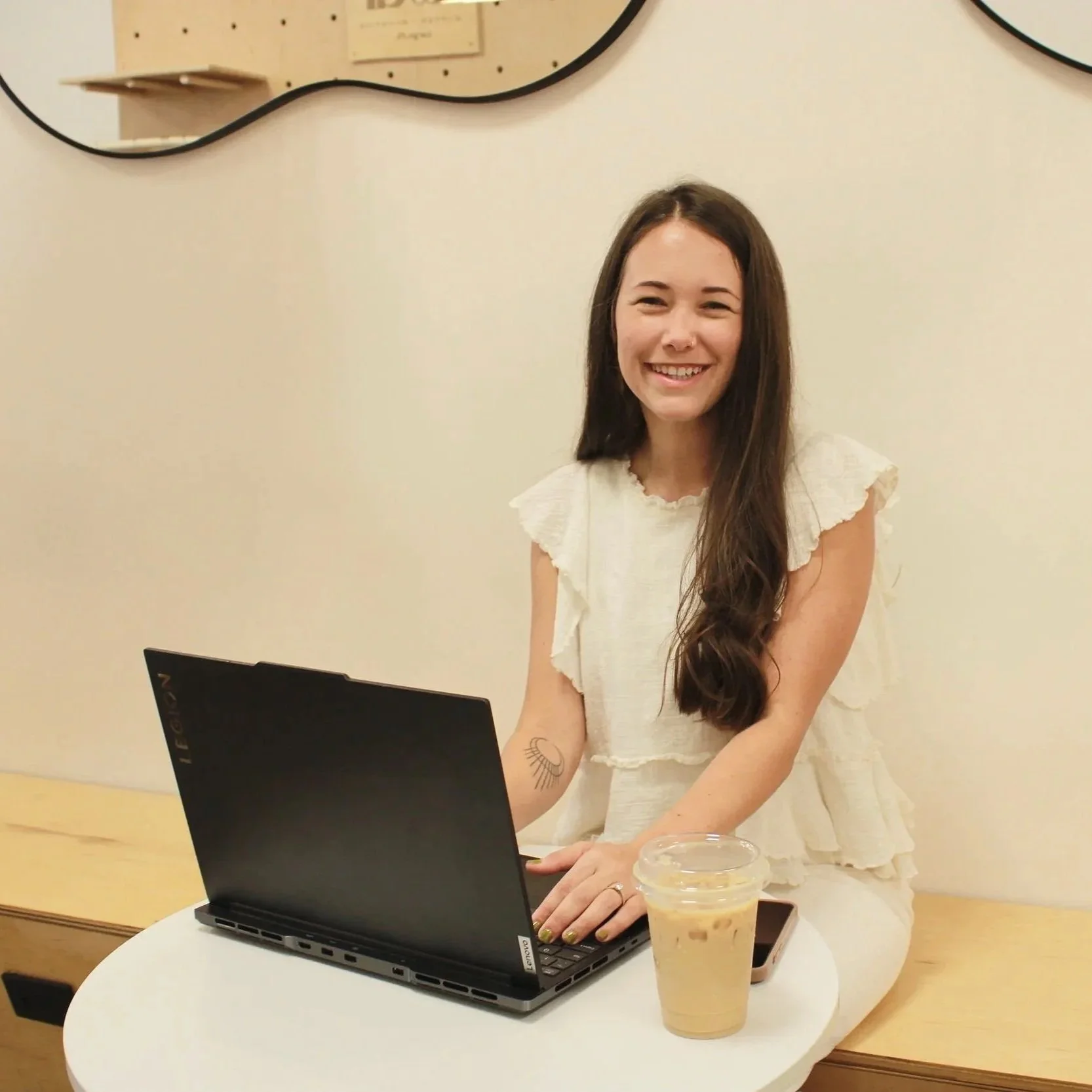A website designer sitting at a neutral minimalist coffee shop in Clearwater, Florida. She is smiling at the camera while typing on her laptop and has an iced latte on the table next to her computer.