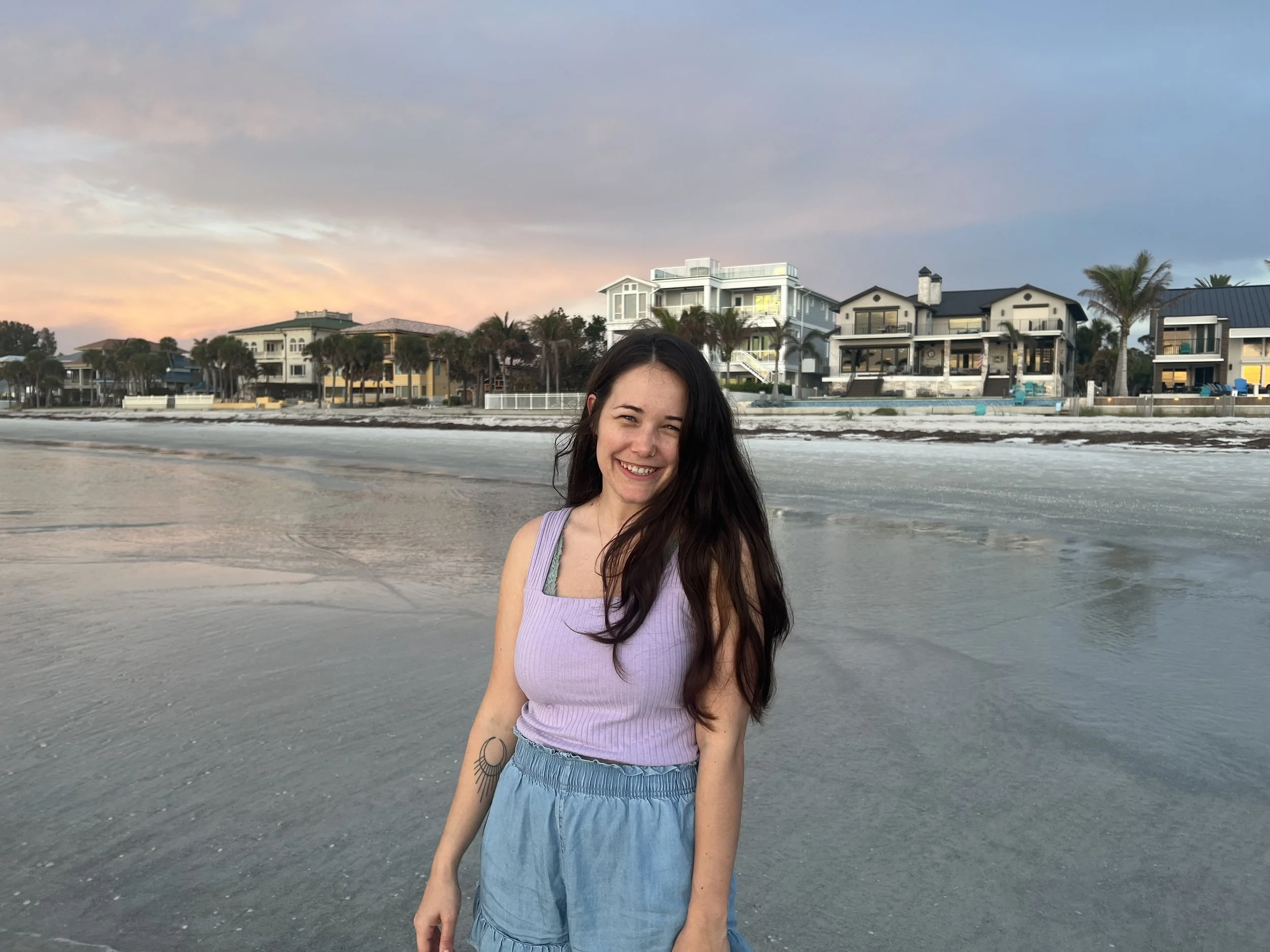 A woman standing and smiling on the beach in the water at sunrise with beach houses and a pink sky behind her.