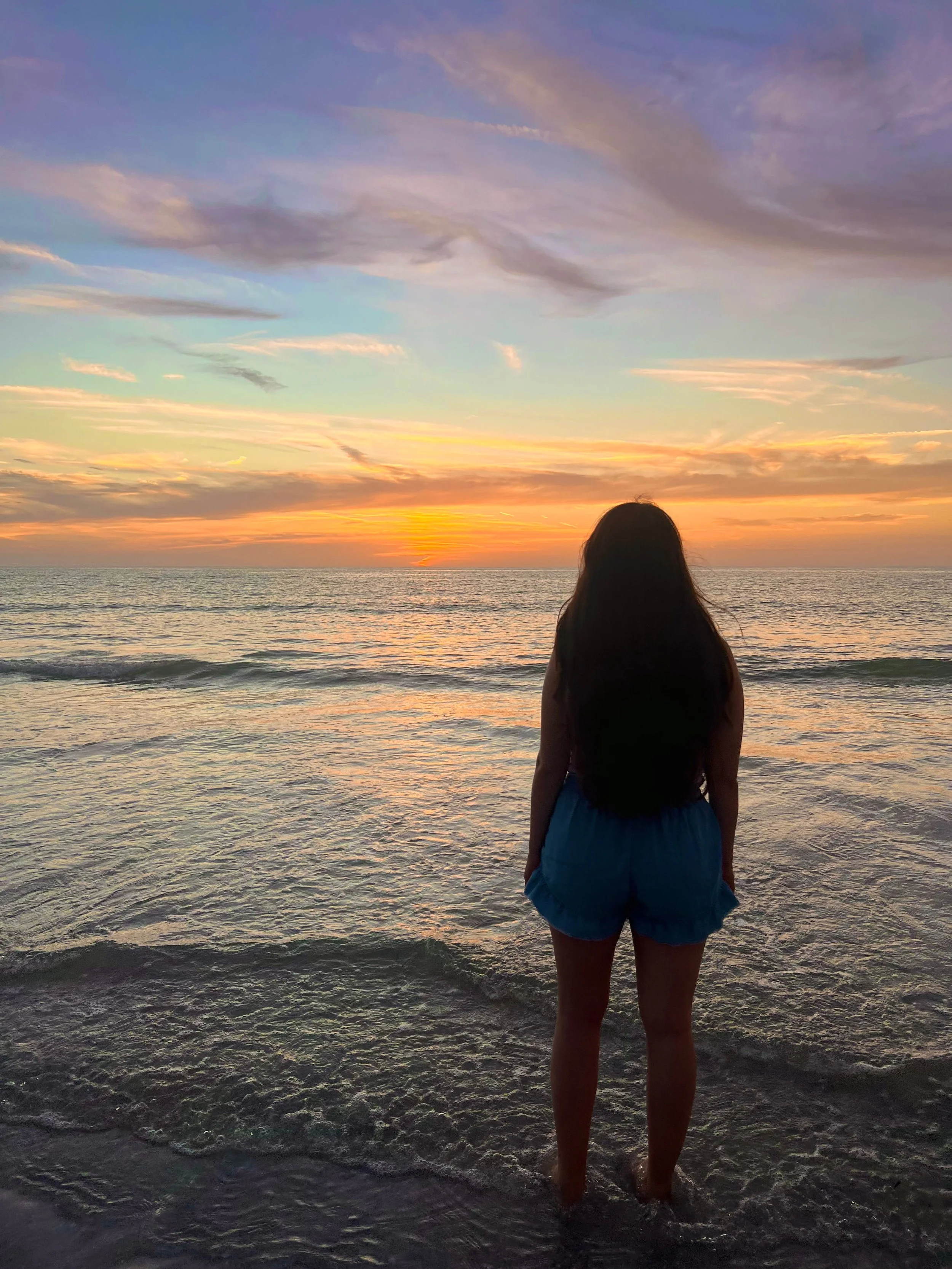 The silhouette of a woman standing on the shore and looking out to the horizon during an orange and blue sunset.