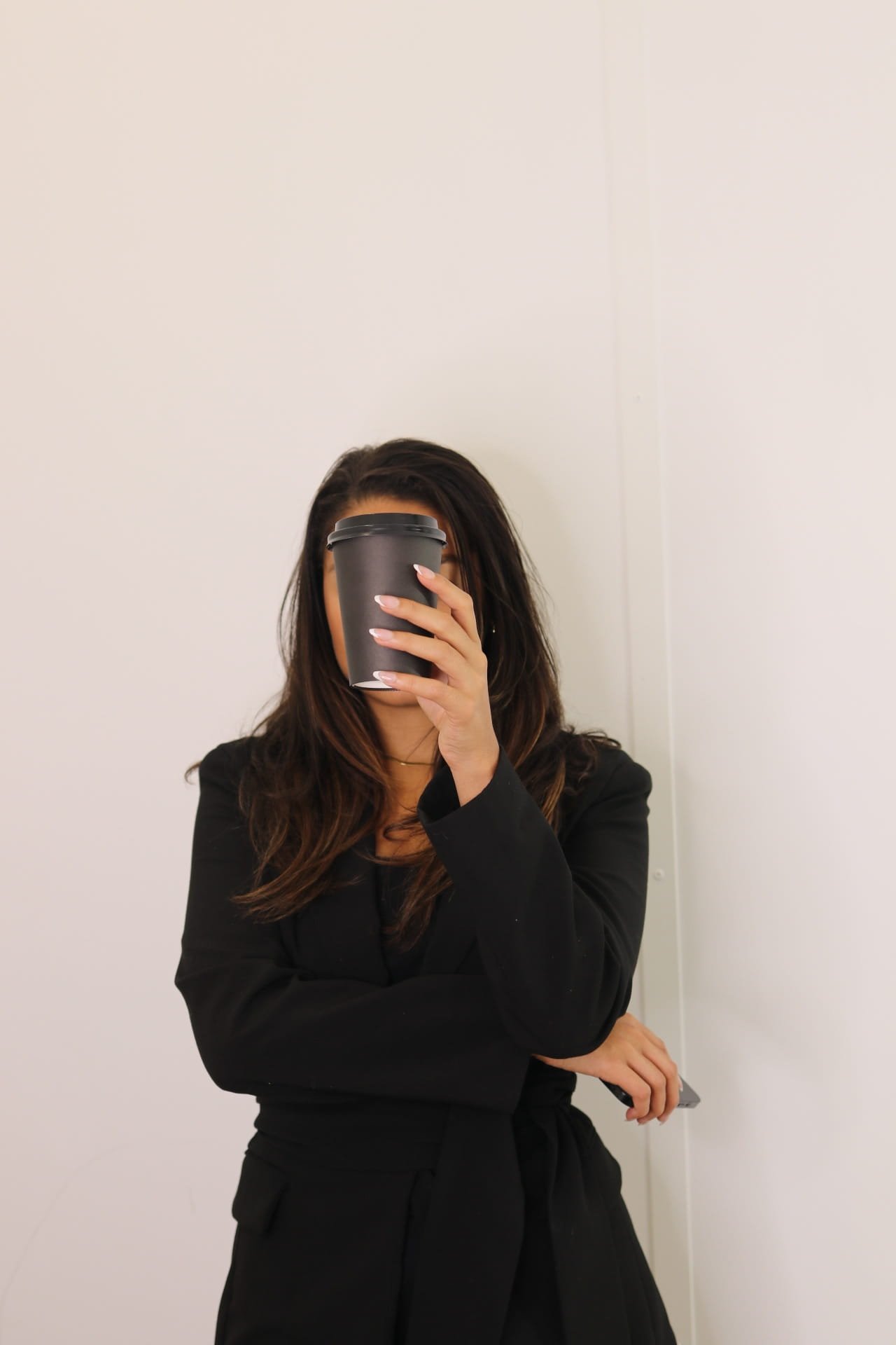 Modern, polished woman wearing a blazer and holding a to-go coffee cup in front of her face while her other arm is crossed under with her phone in her hand.