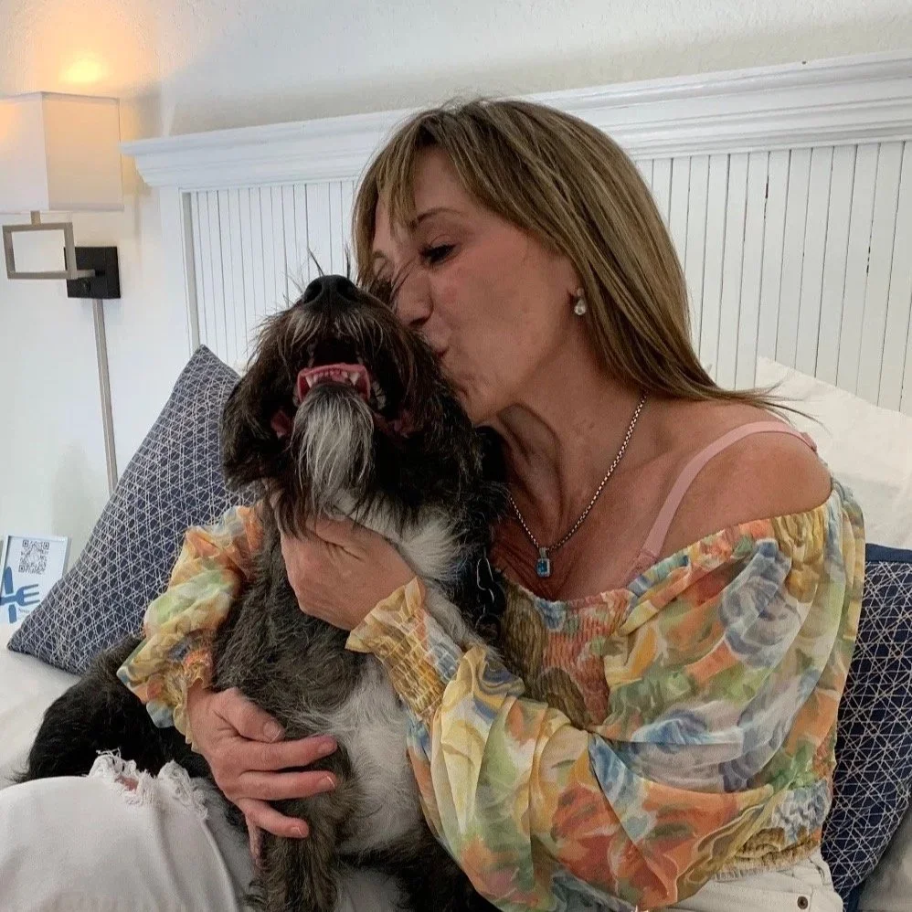 Woman kissing dog on the face while holding it in her arms on bed, bedroom setting.
