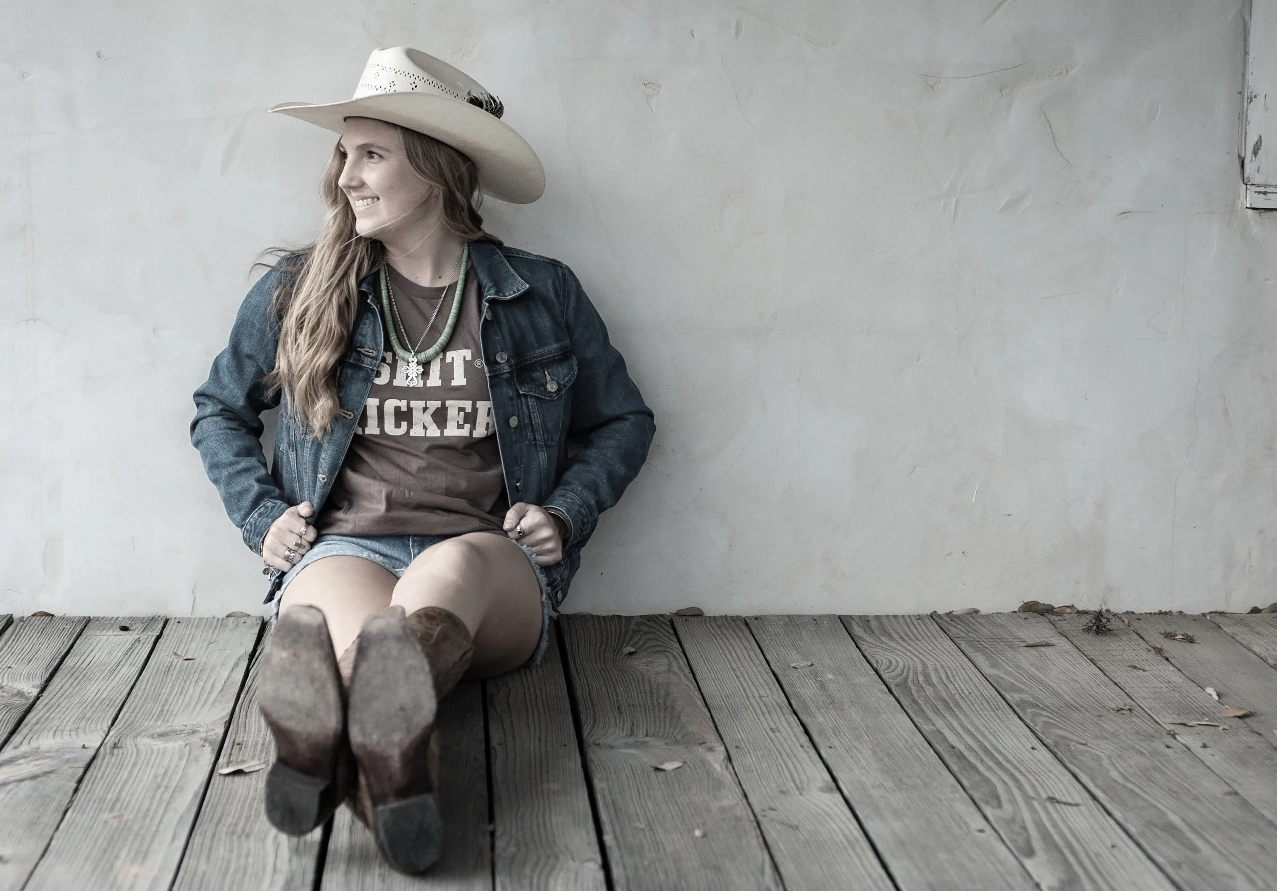 Young woman with long hair, wearing a cowboy hat, denim jacket, and casual clothes, sitting on a wooden deck against a plain wall, smiling and looking to the side.