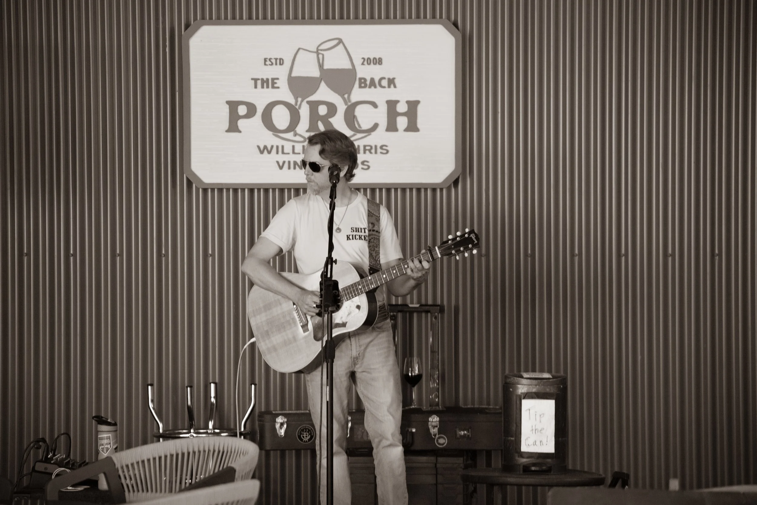 A man with long hair, wearing sunglasses and a t-shirt, playing an acoustic guitar. He is standing in front of a sign that reads 'The Porch' and has a drink in a glass on a nearby table.