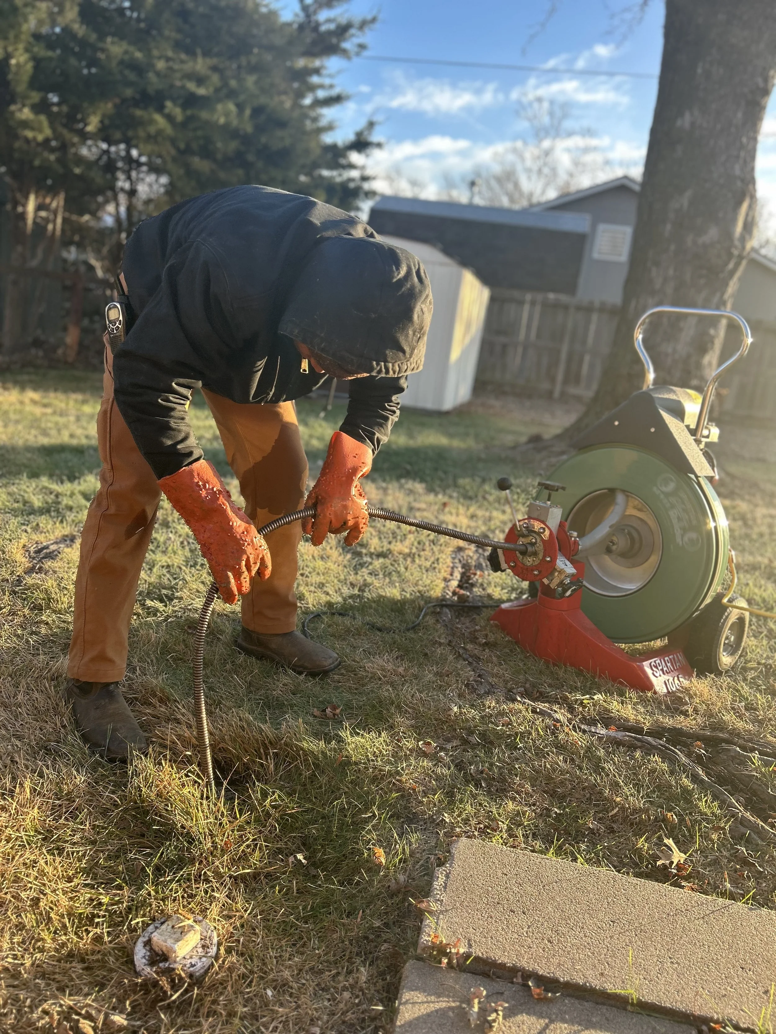 Man wearing orange gloves, black hoodie, and brown pants working on a yellow saw cutting concrete in a backyard with trees and a shed, sunlight casting shadows.