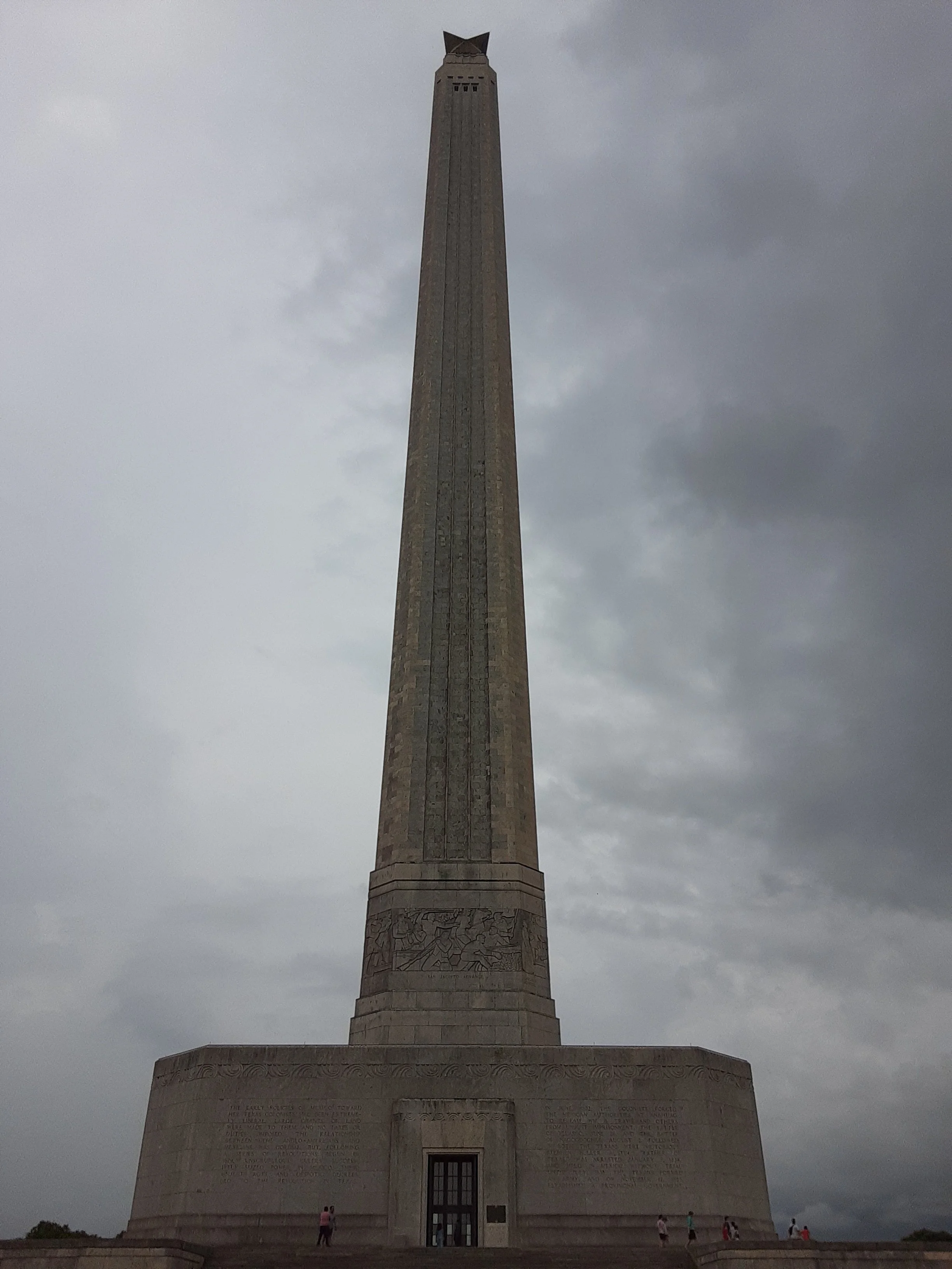 A tall stone tower with a carved base and inscription plaques at the bottom, topped with a structure resembling an owl, under cloudy skies with park visitors walking around.