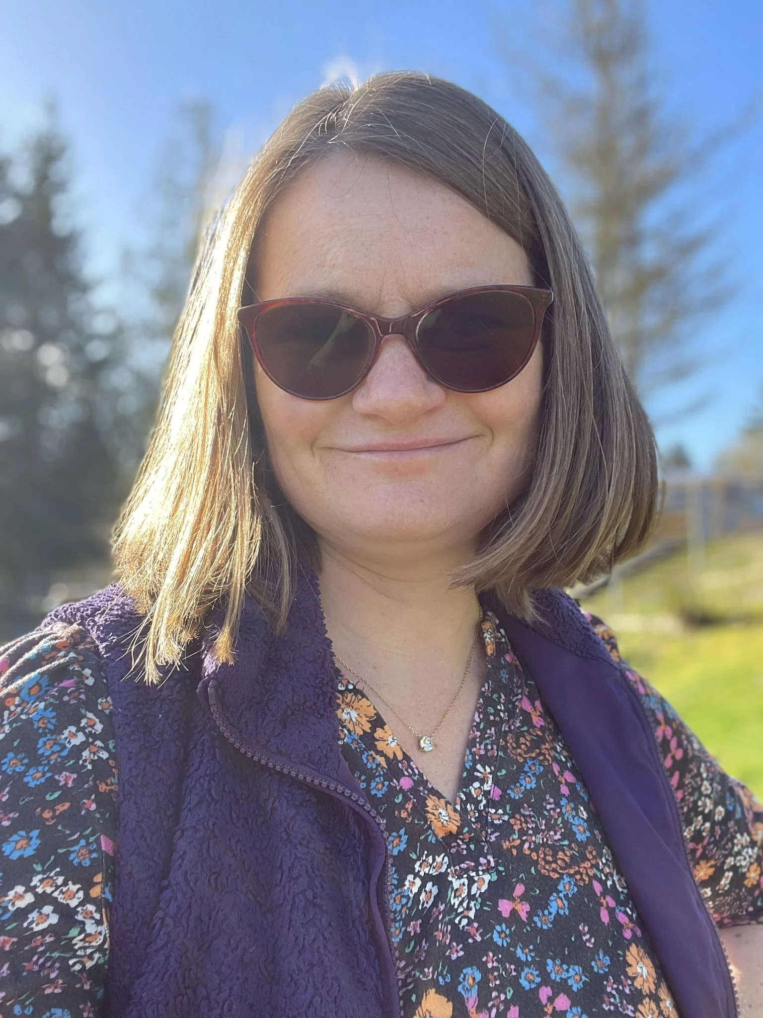 A therapist, Lynn Jones, wearing sunglasses and a floral blouse, a purple vest standing outdoors on a sunny day. Showing who you would be meeting with if you chose to start therapy at Root of Life Counseling