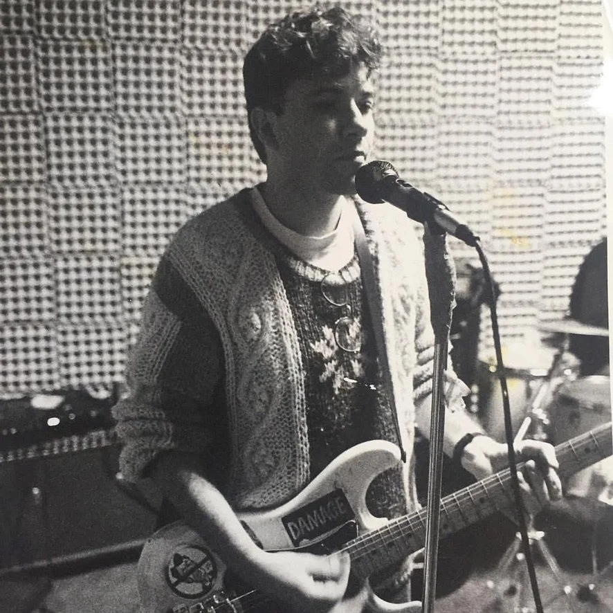Black and white photo of a young man singing into a microphone while playing an electric guitar in a music rehearsal space.