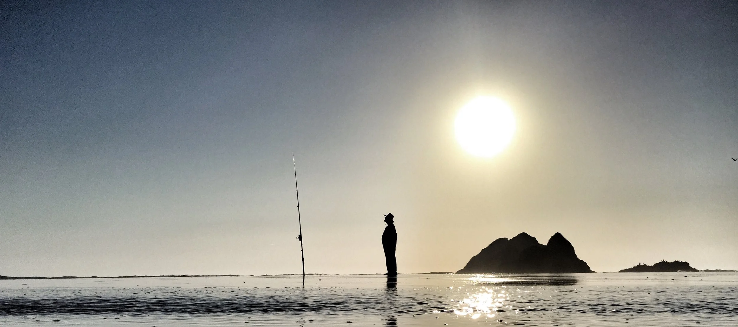 Silhouette of a person fishing on the beach at sunset with rocks and a small island in the background.