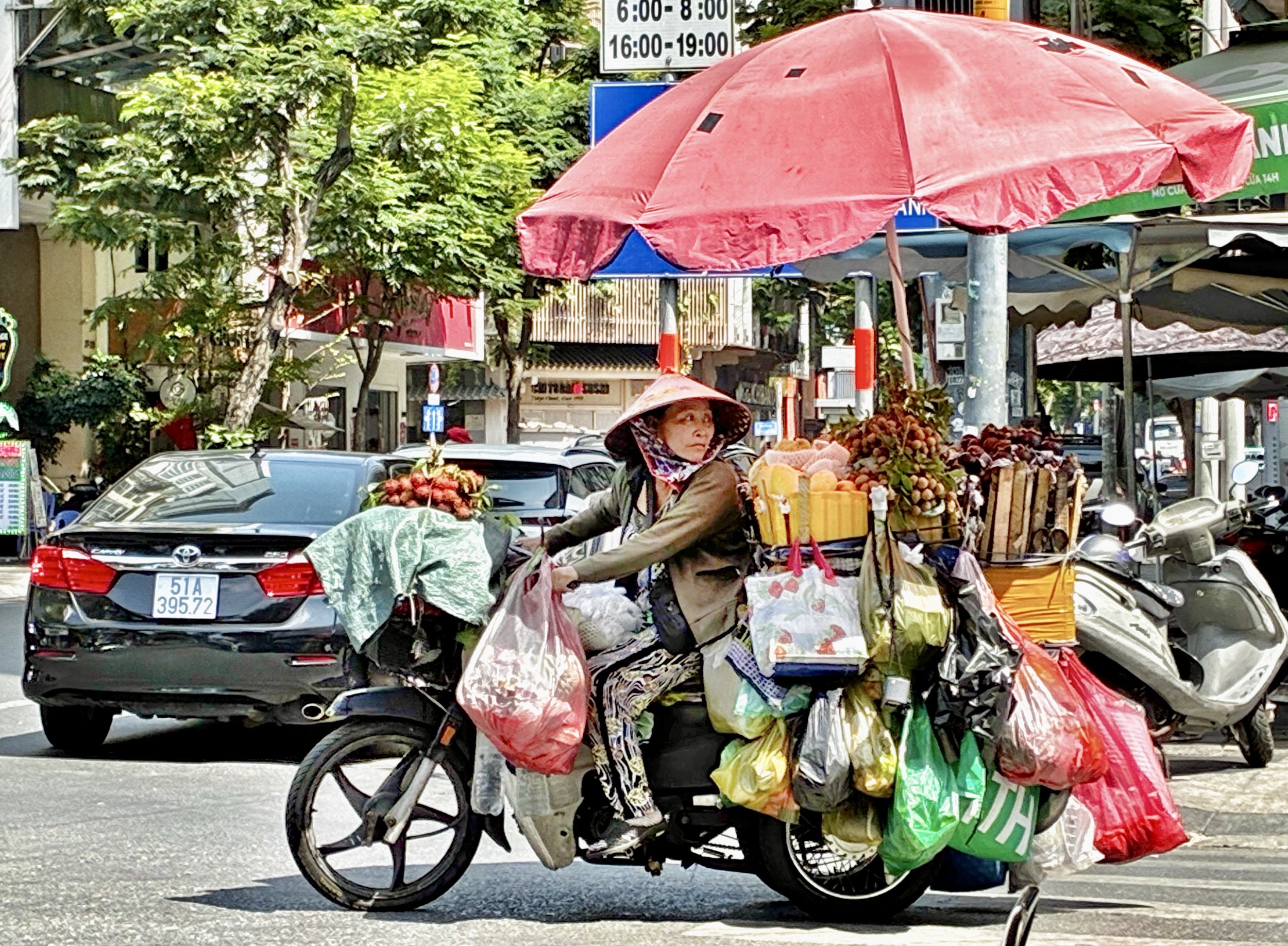 A woman riding a bicycle on a city street, covered with numerous bags and boxes filled with fruits and other goods, with a red umbrella over her head, cars parked on the side of the road, and trees and signage in the background.