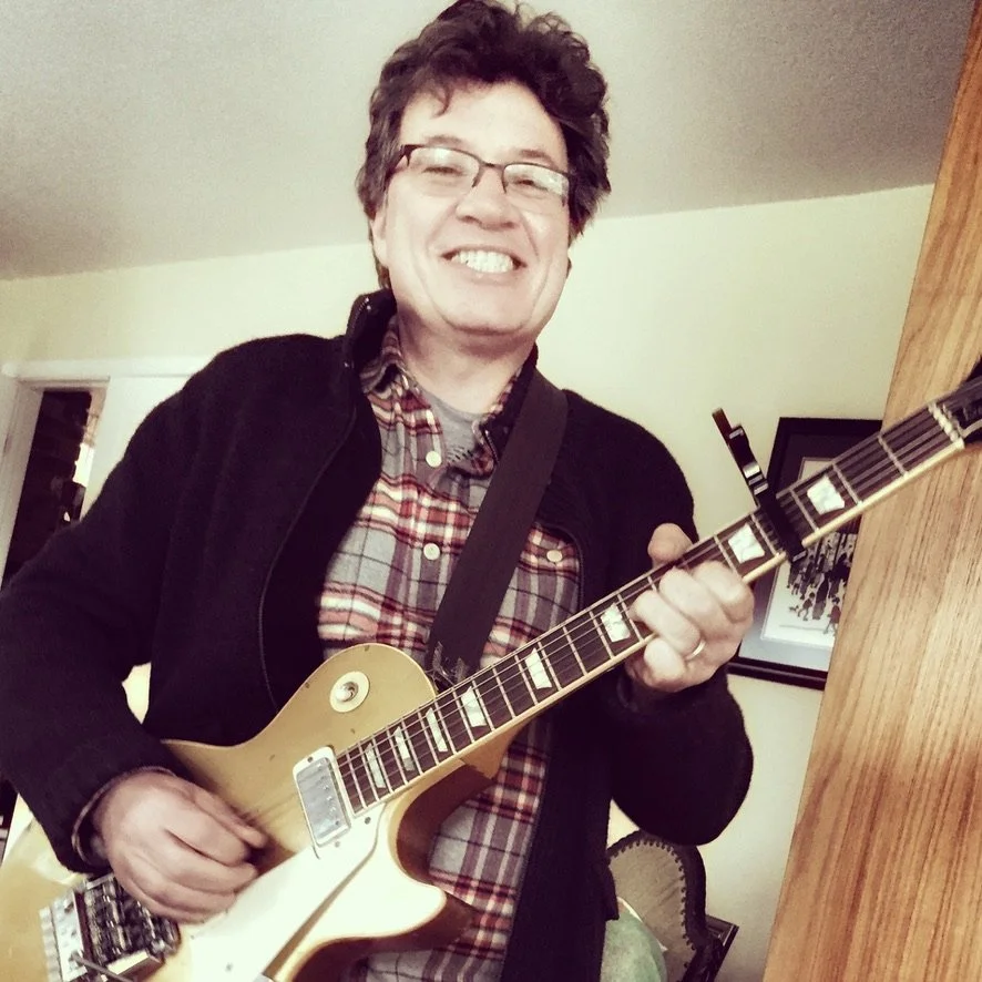 A man wearing glasses and a plaid shirt, smiling and playing a yellow electric guitar indoors.
