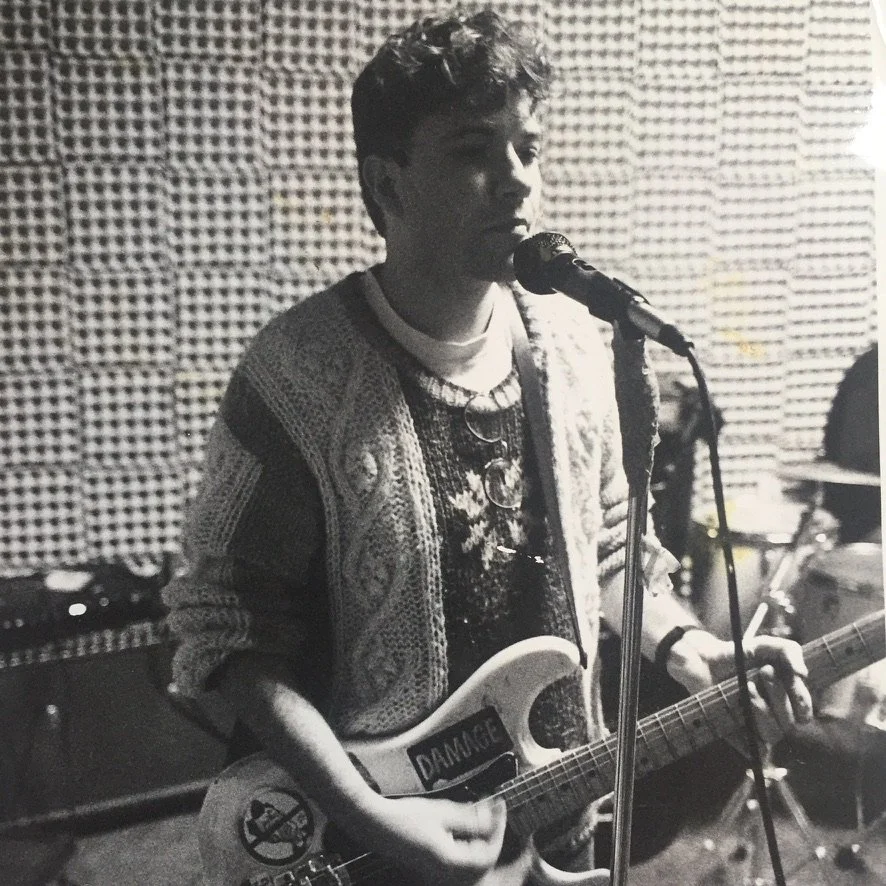 A young man with sunglasses hanging from his shirt plays an electric guitar and sings into a microphone in a music studio with soundproofing foam on the walls.