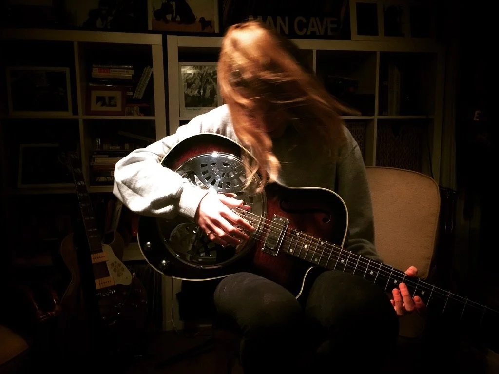 Person with long hair playing an electric guitar in a dimly lit room with shelves and books in the background.