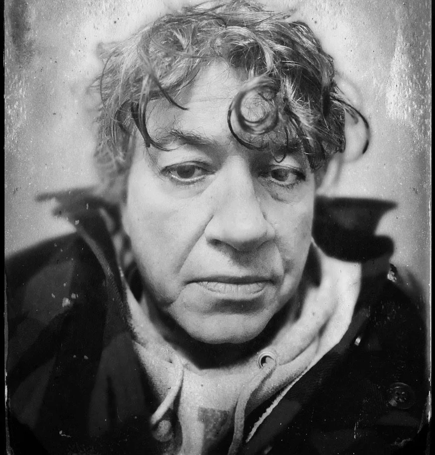 A black and white close-up photo of a young man with curly hair and a serious expression, wearing a hoodie and a jacket.