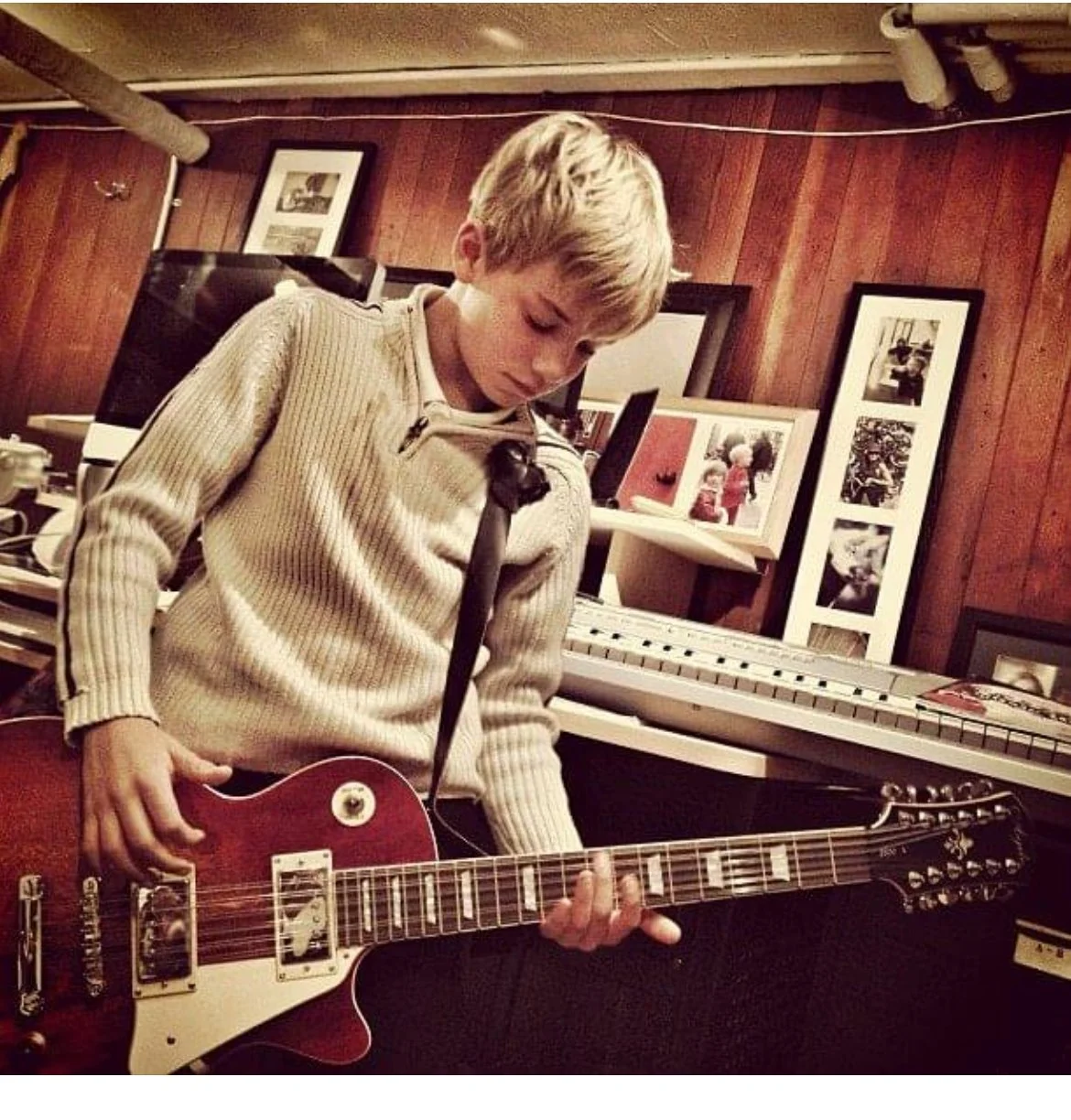 A young boy playing a red electric guitar in a room with wooden walls, surrounded by framed photos and a keyboard in the background.