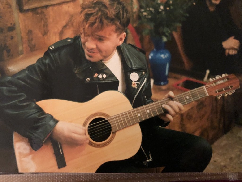 A man playing acoustic guitar in a cozy indoor setting with wooden walls and a blue vase in the background.