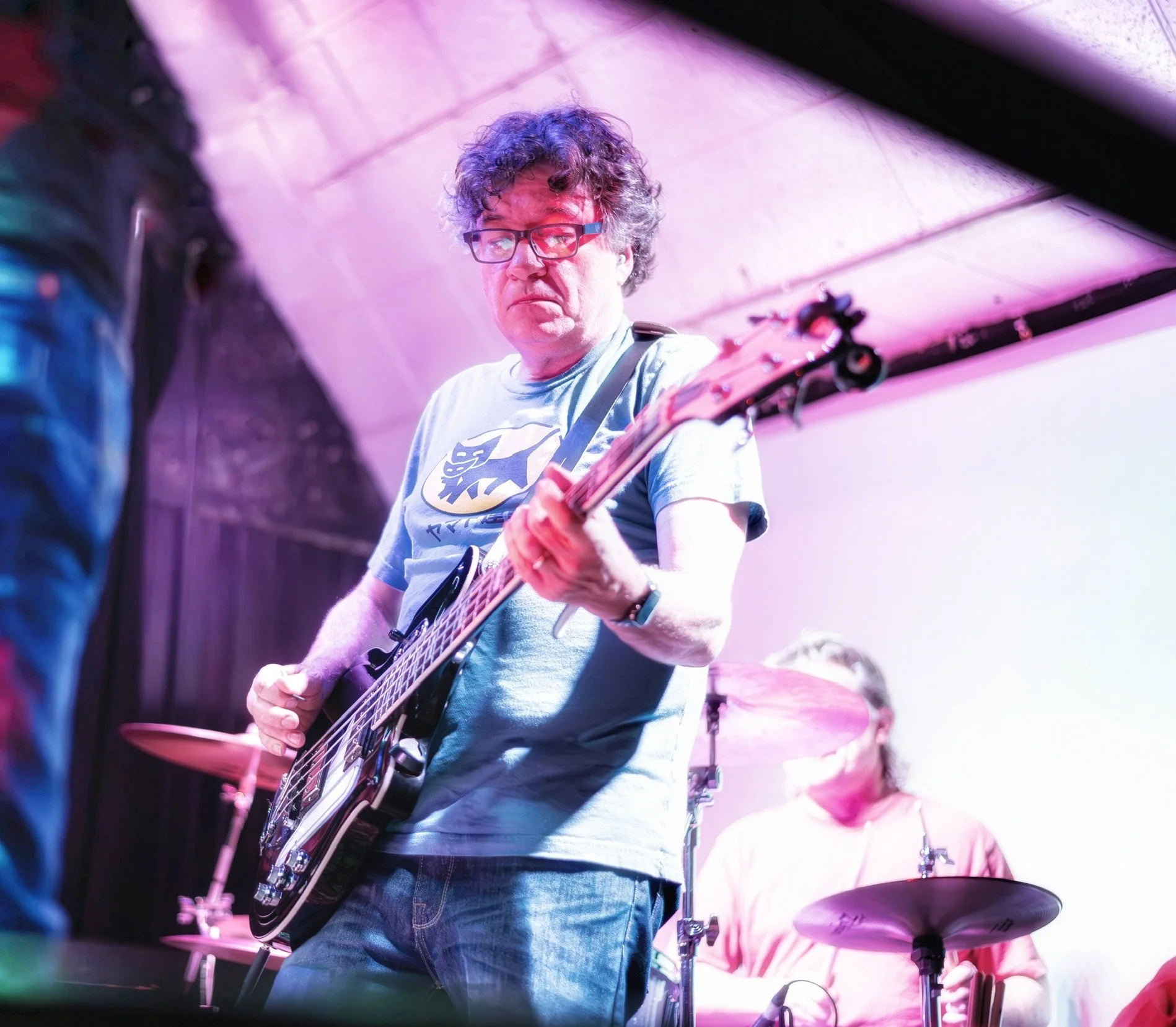 A man with glasses and curly hair playing an electric guitar on stage with colorful lighting, and a drummer playing a drum set in the background.