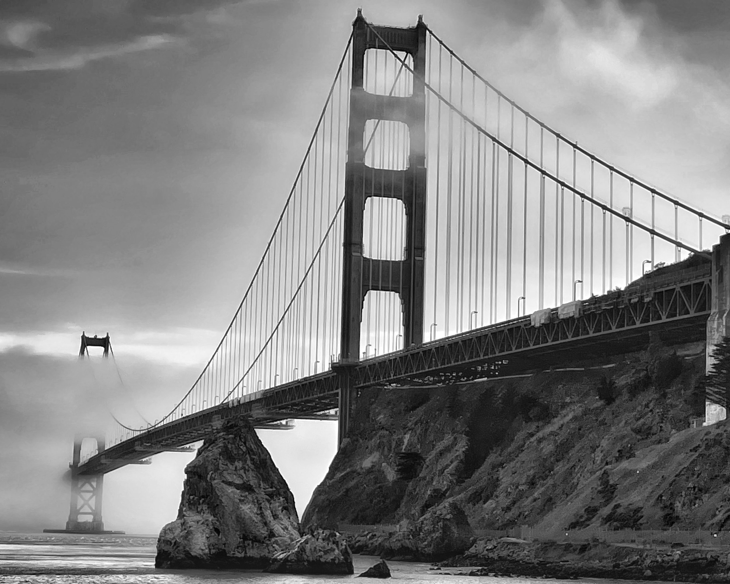 Black and white photo of the Golden Gate Bridge with a large rock formation and water in the foreground.