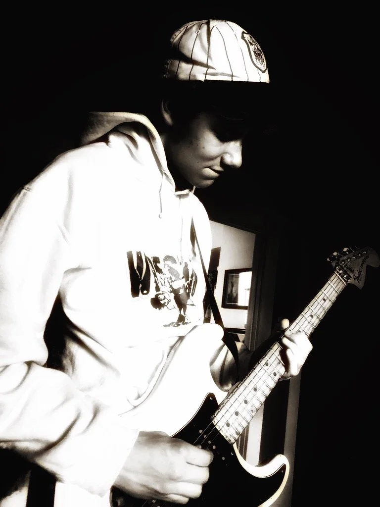 A young person playing an electric guitar, wearing a baseball cap and sunglasses, in a black and white photo.