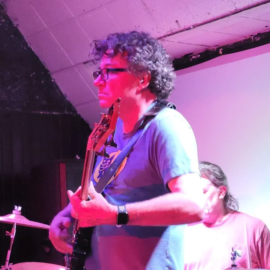 A person with curly hair and glasses playing an electric guitar on stage, with another person partially visible playing drums in the background, under colorful stage lights.