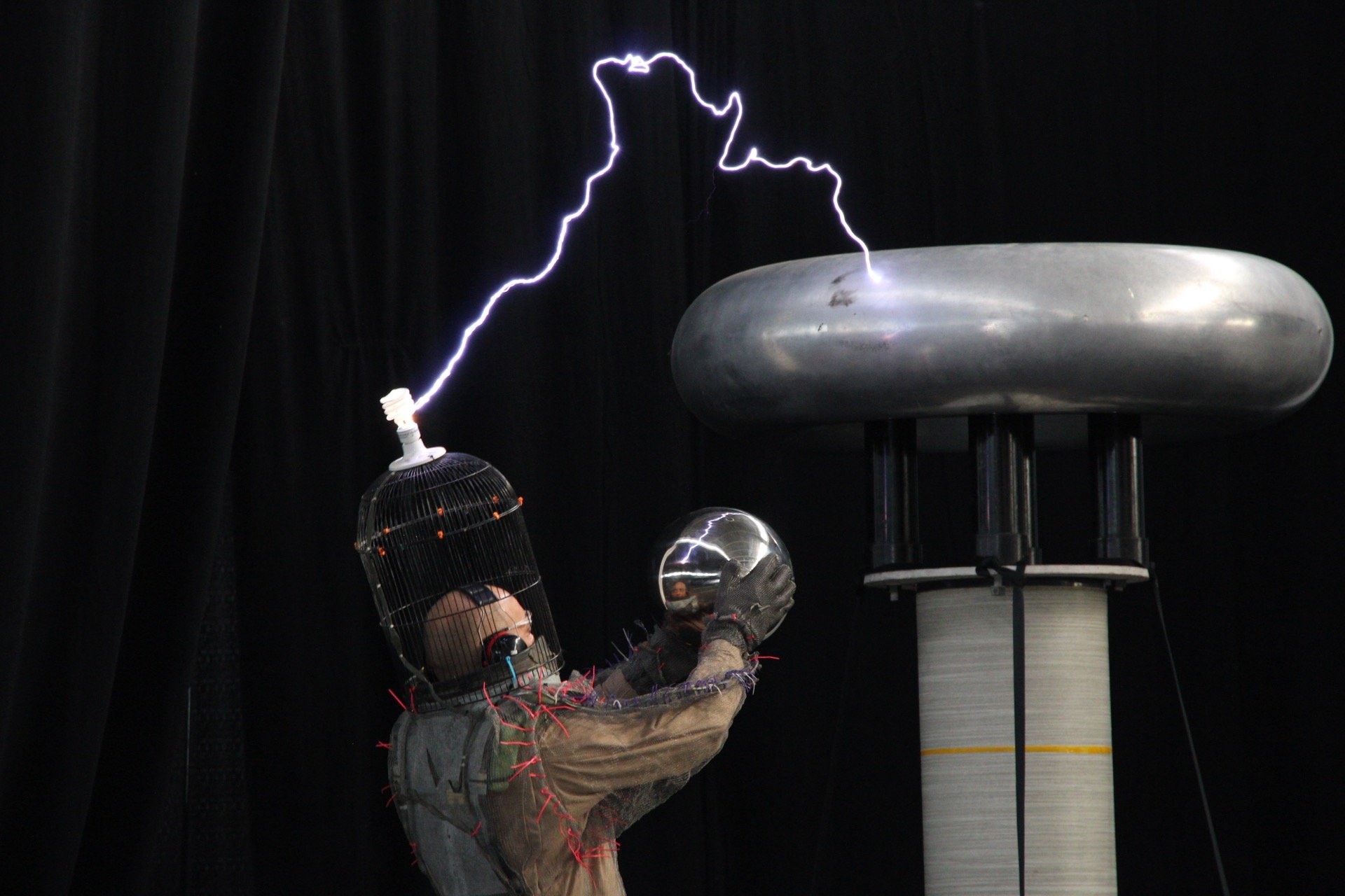 A person wearing protective gear and a cage on their head is conducting a static electricity experiment, generating lightning between a metal sphere and a lightning rod.
