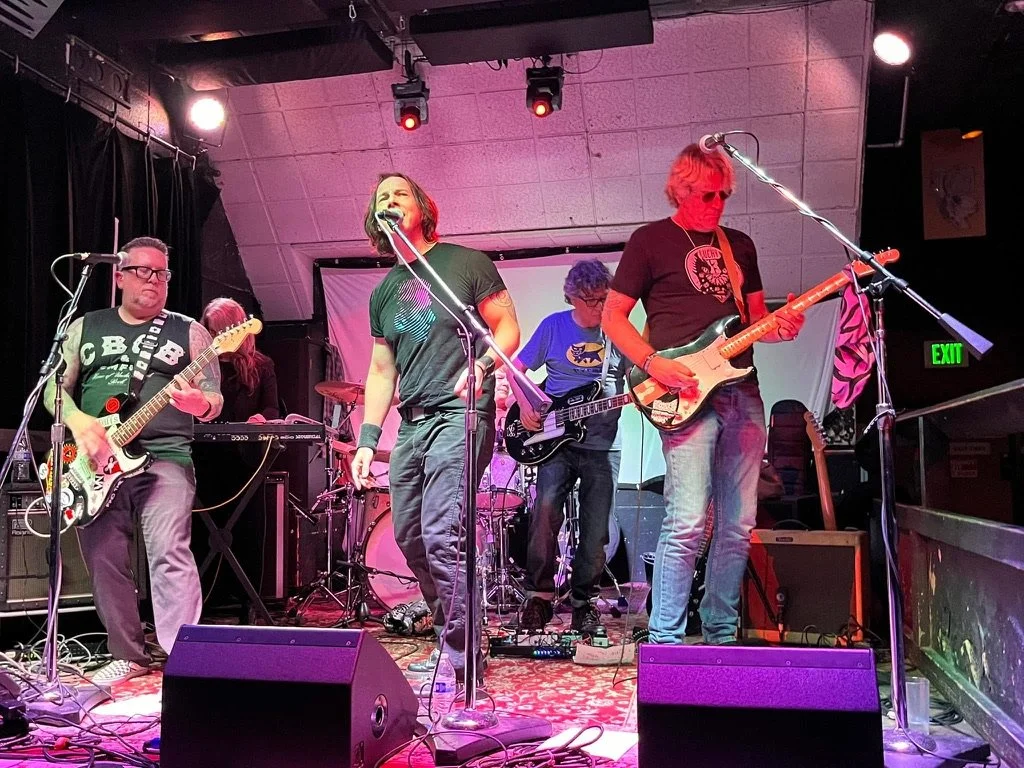 A five-piece band performing on stage in a nightclub. The lead singer, a woman, stands center with a microphone. Guitarists and a keyboard player are on either side, with a drum set behind them. Stage lighting is visible overhead and a patterned carp
