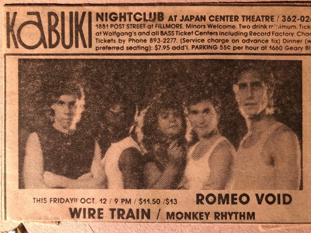 A black and white photo of five people, likely band members, posing together. The background is dark with some outdoor elements visible. They are dressed in 1980s style clothing.