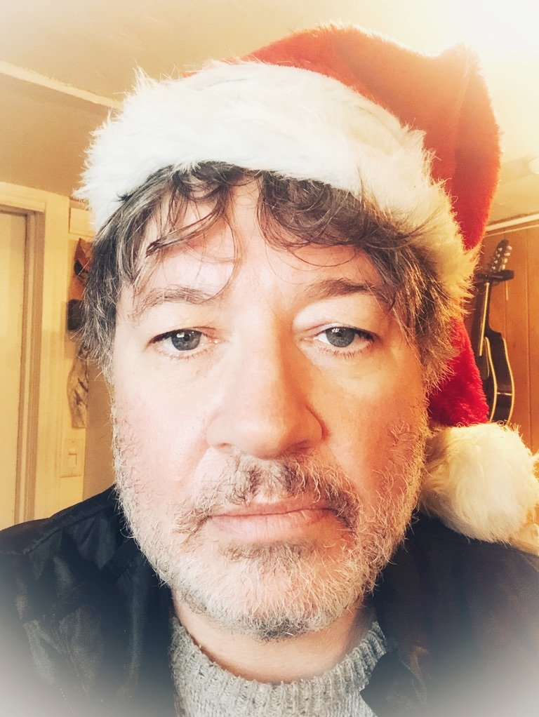 A man wearing a Santa hat with a white faux fur trim and pompom, taking a close-up selfie indoors, with guitars hanging on the wall in the background.
