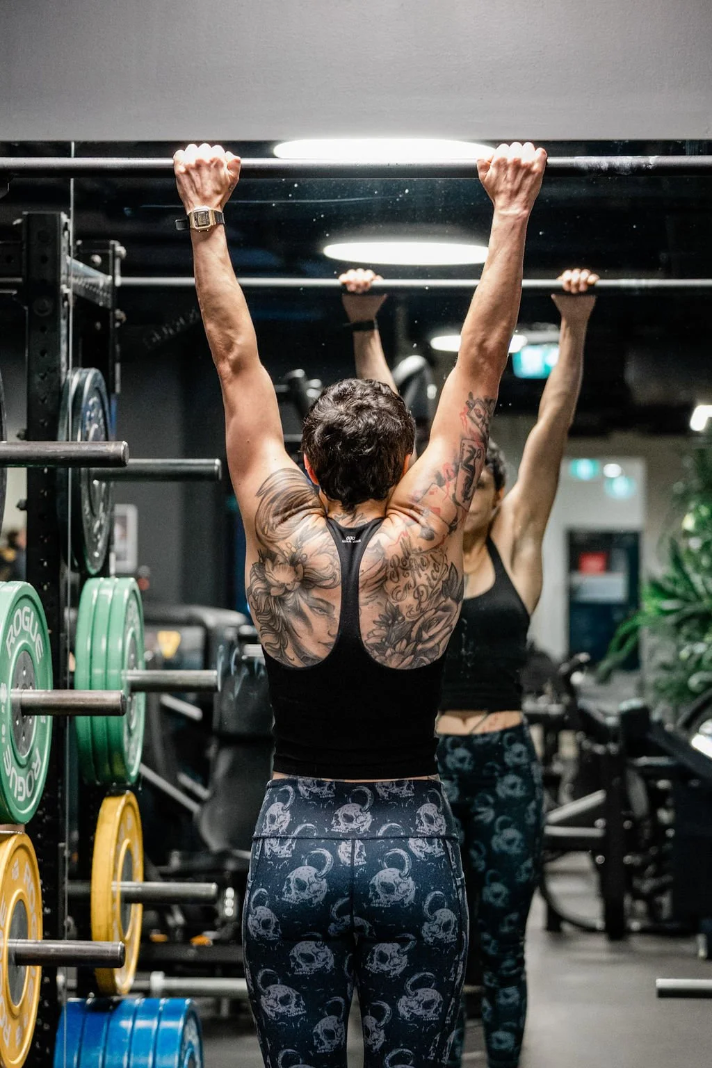 A woman with tattoos on her back is doing a pull-up in a gym, with another woman in workout clothes reflected in a mirror behind her.
