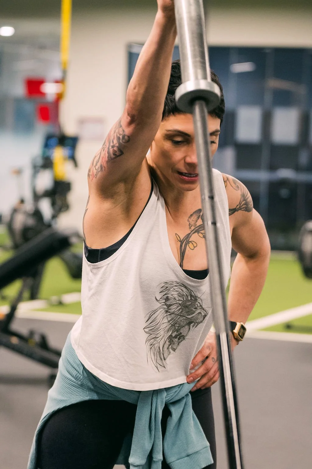 A woman with tattoos on her arms and chest, wearing a white tank top with a lion illustration, exercises with a barbell in a gym.
