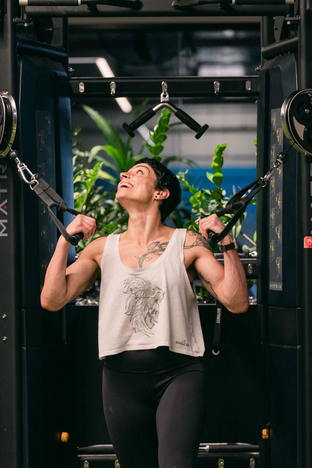 A woman with short dark hair and tattoos on her arms and chest, smiling and lifting weights in a gym surrounded by green plants.