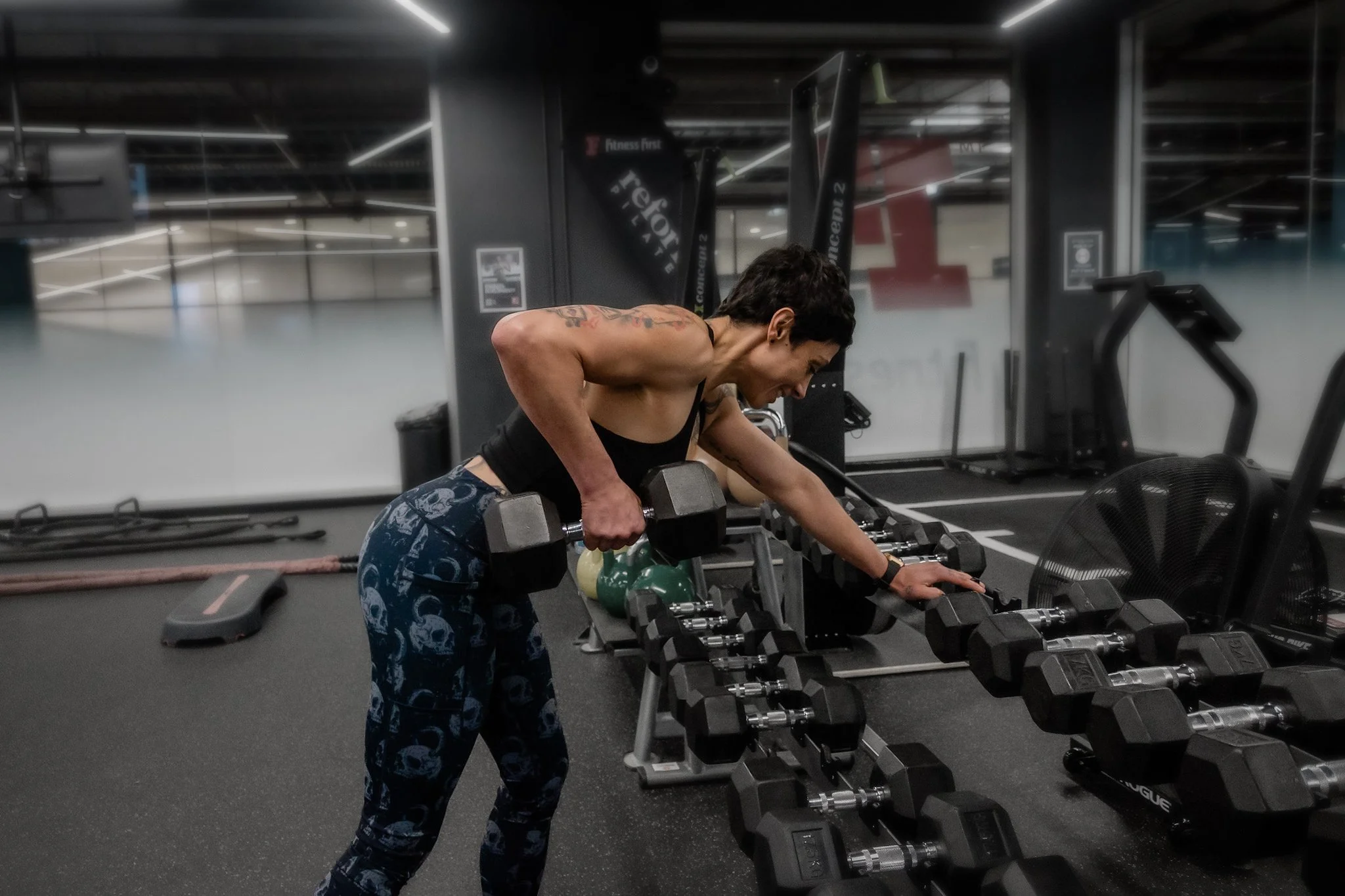 A woman working out in a gym, performing a bent-over dumbbell row exercise. She is wearing a black tank top and blue patterned leggings, with short dark hair and tattoos on her arms. The gym has a black workout area with various equipment in the background, including dumbbells on racks.