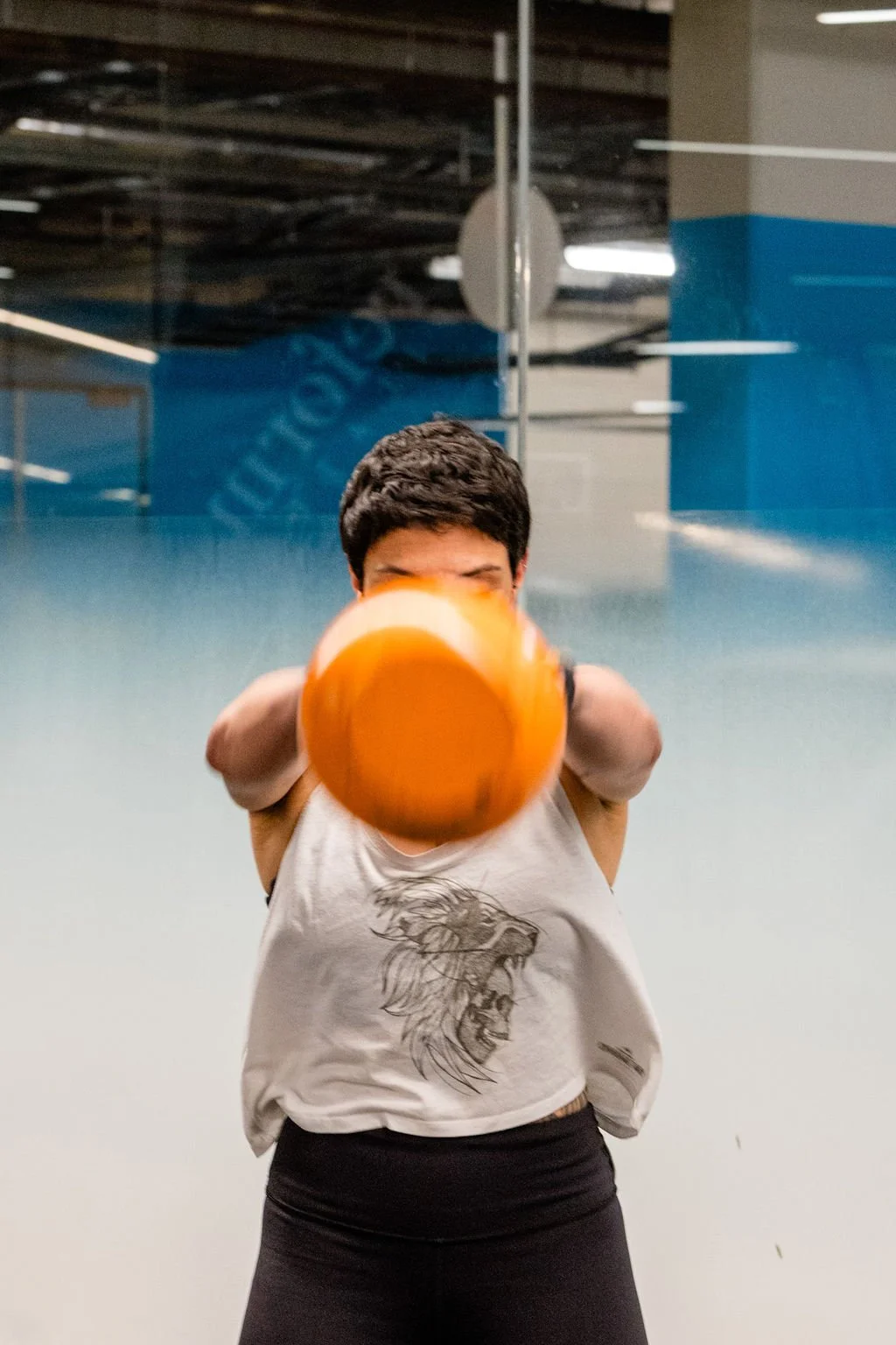 Person holding a large orange kettlebell towards the camera in an indoor gym.