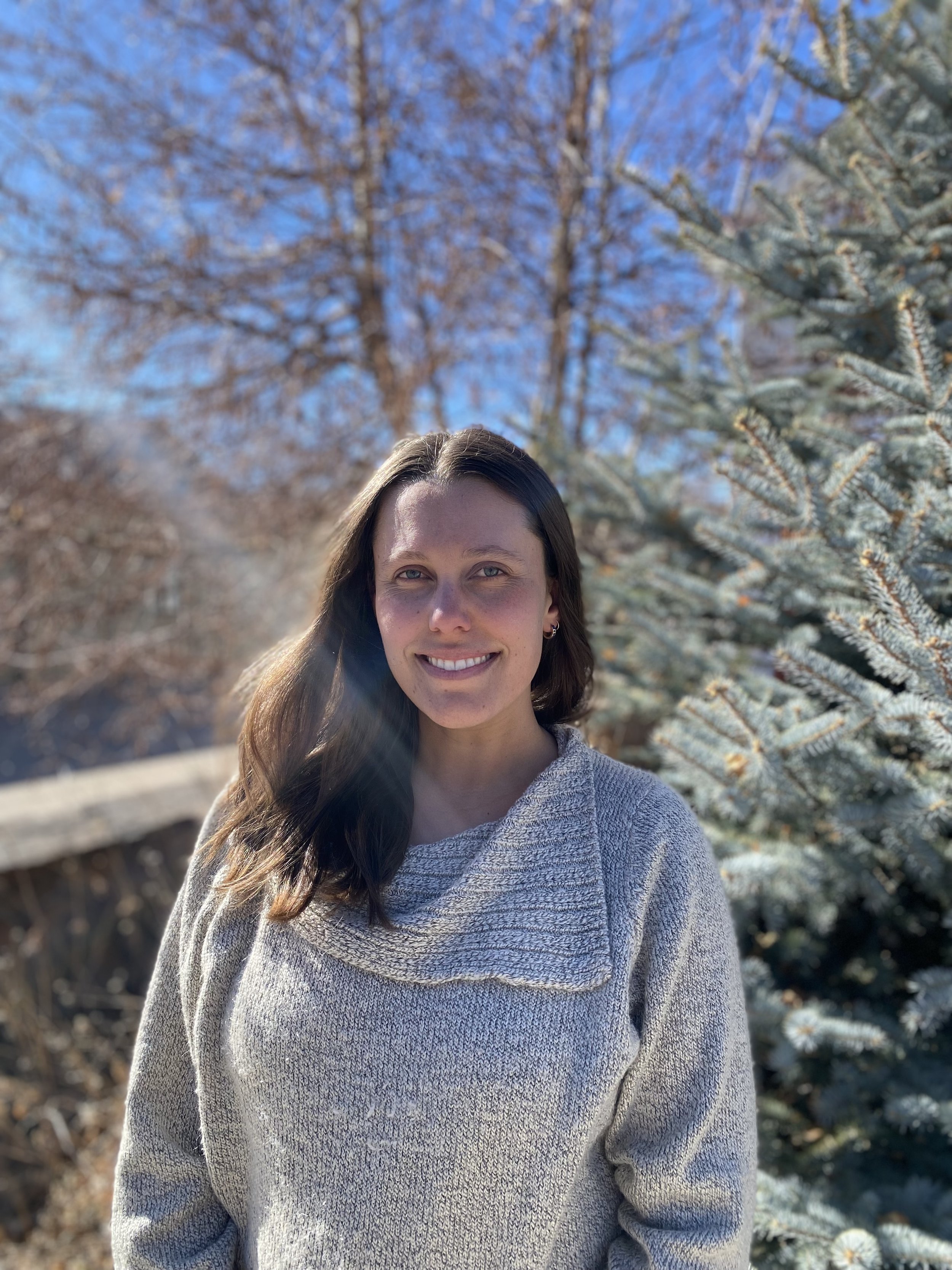 A woman with long brown hair and light skin, smiling outdoors in front of a snow-covered pine tree and trees with bare branches, wearing a light gray sweater.