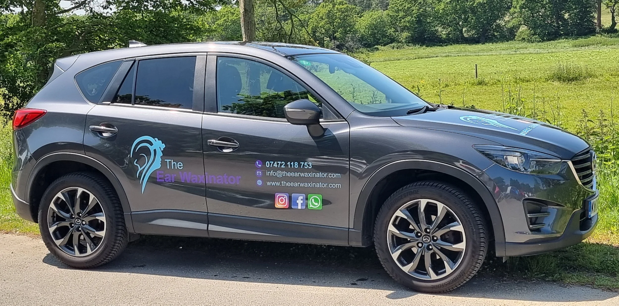 A gray SUV parked on the side of a road with a green grassy field and trees in the background. The vehicle has branding for 'The Ear Waxinator' with contact information and social media icons on the side.