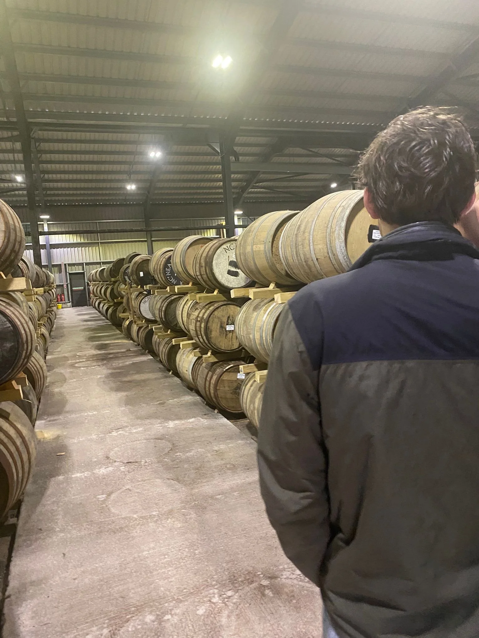 Inside a warehouse with metal roofing and shelves holding wooden barrels, with a man in a black jacket facing away and taking a photo of the barrels.