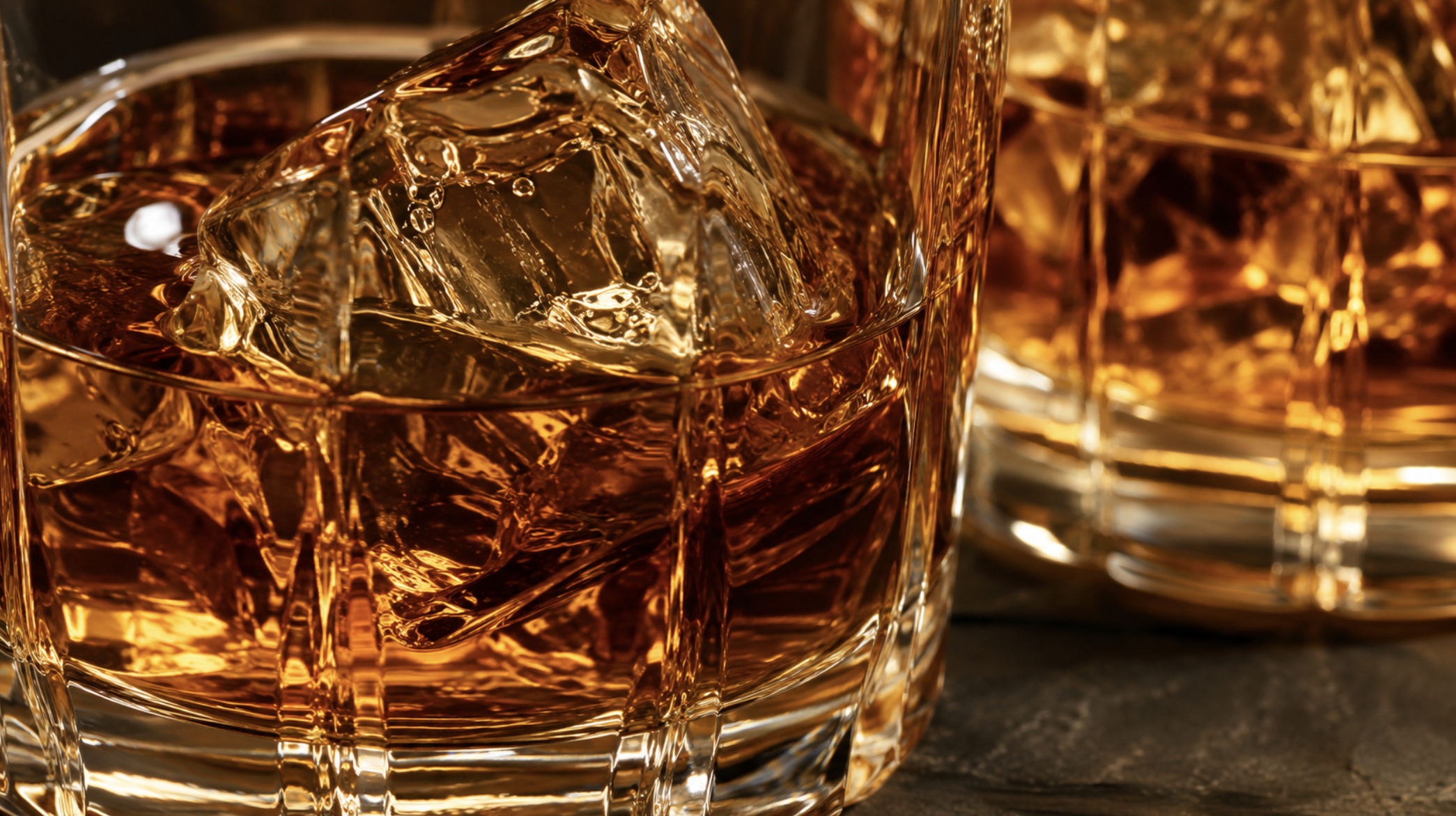 Close-up of a glass of whiskey with ice cubes, reflecting light, with more glasses of whiskey in the background.