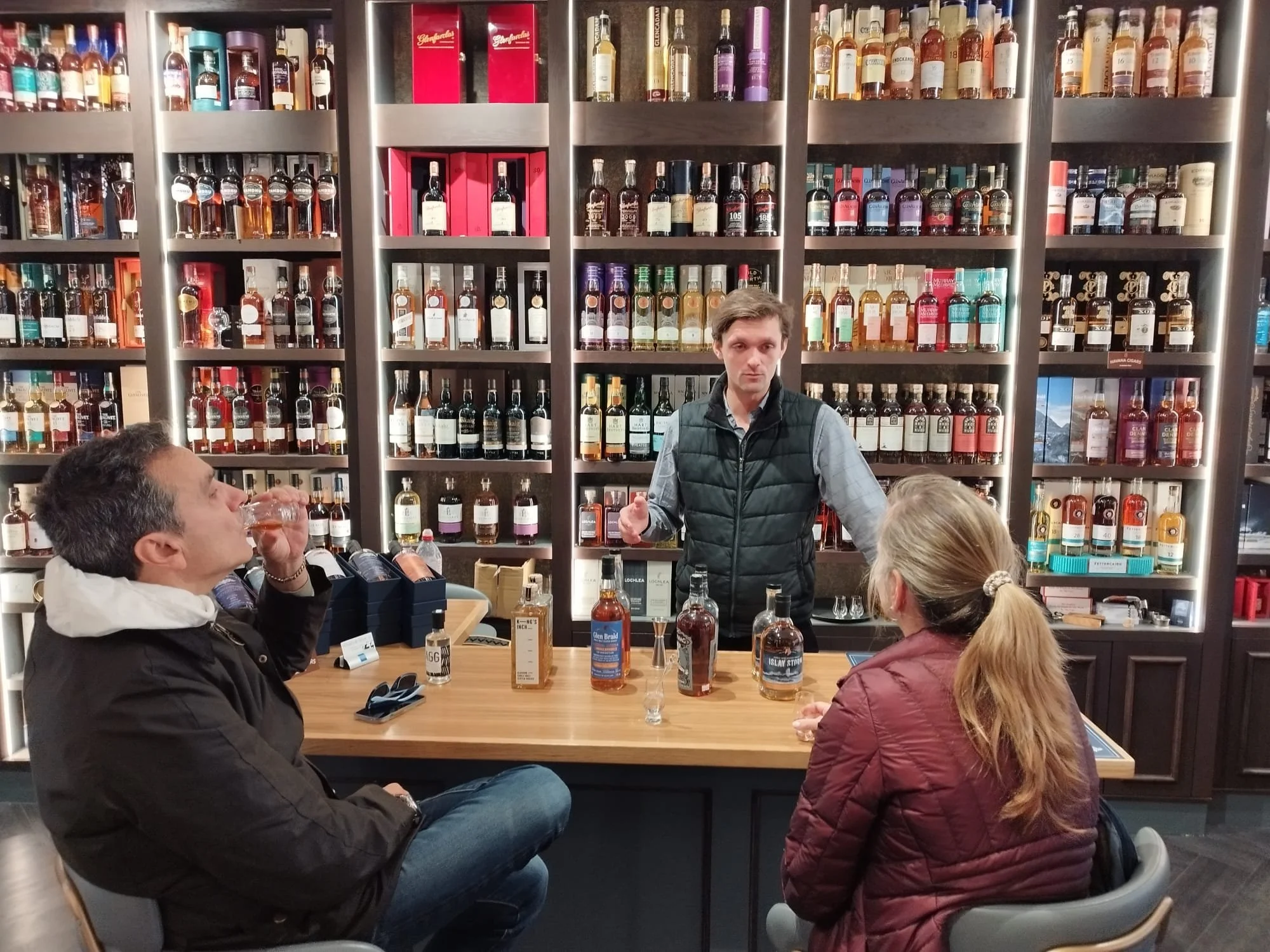 People tasting and discussing whiskey at a tasting event in front of shelves filled with various whiskey bottles.