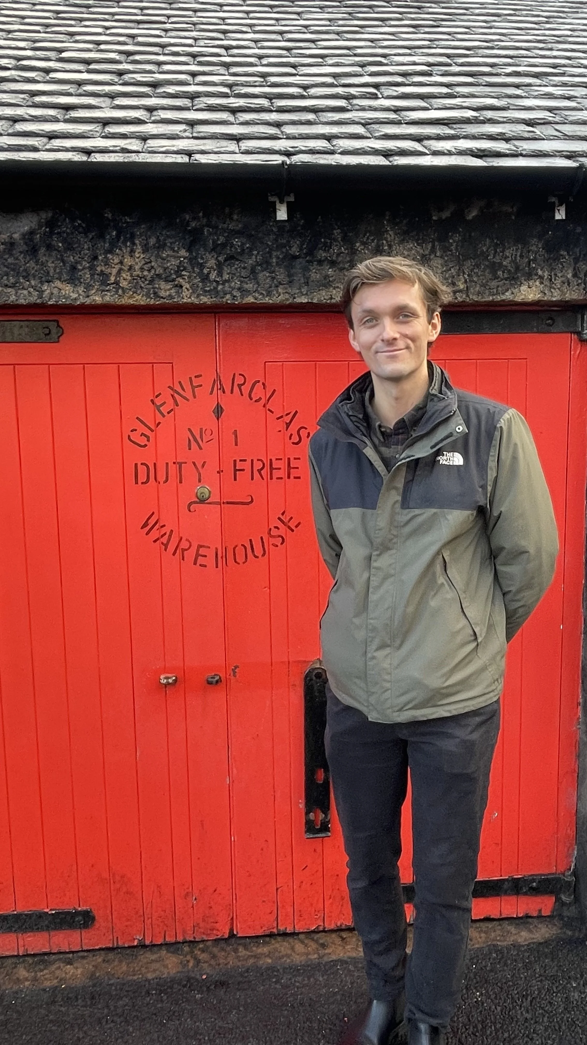 A man standing in front of a bright red door with a sign that reads, 'Glenfarg Lass Duty Free Wharehouse.'