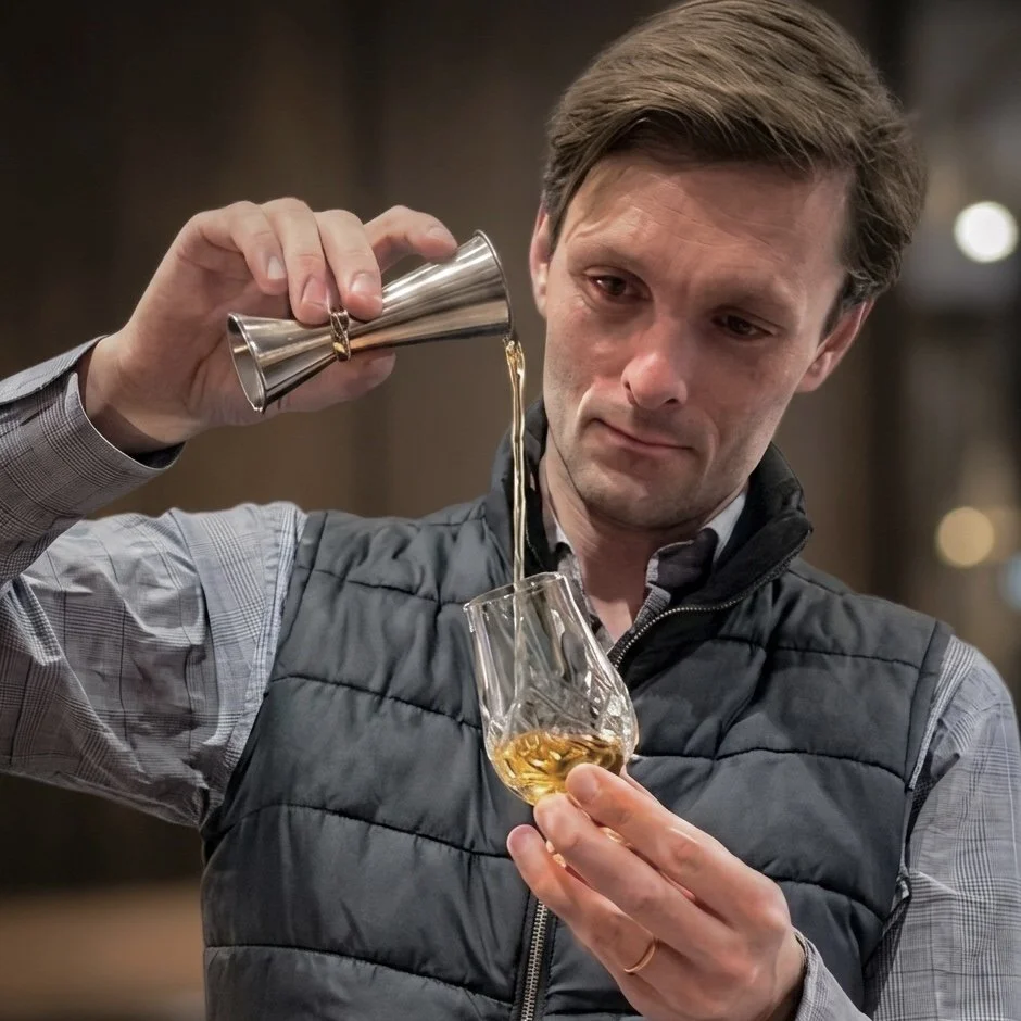 A man pouring whiskey into a glass from a metal jigger in a dimly lit setting.