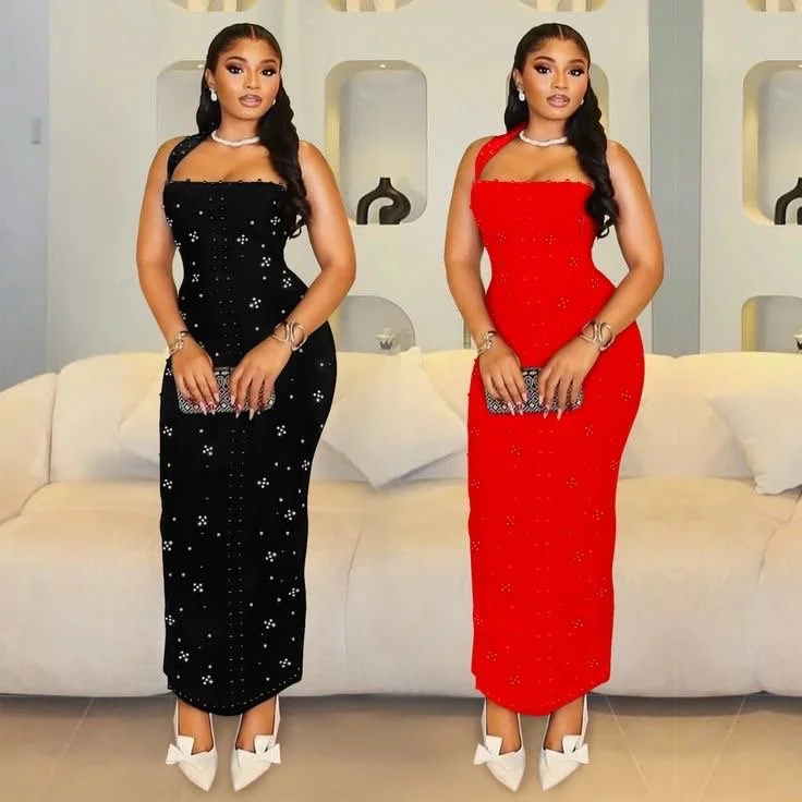 Two women stand side by side in elegant evening dresses, one in black and the other in red, holding small handbags, in a modern indoor setting with white couches and decorative shelves in the background.