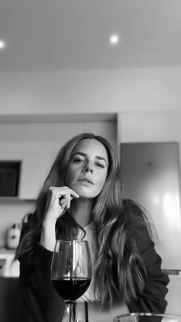 Retrato en blanco y negro de una mujer con cabello largo y suelto, que mira pensativa a la cámara, con un vaso de vino tinto frente a ella en un ambiente de interior moderno.