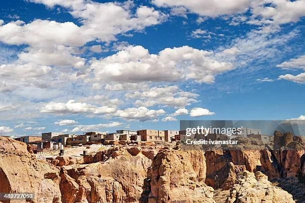 A desert landscape with rocky formations and a few small buildings or structures in the background under a partly cloudy sky.
