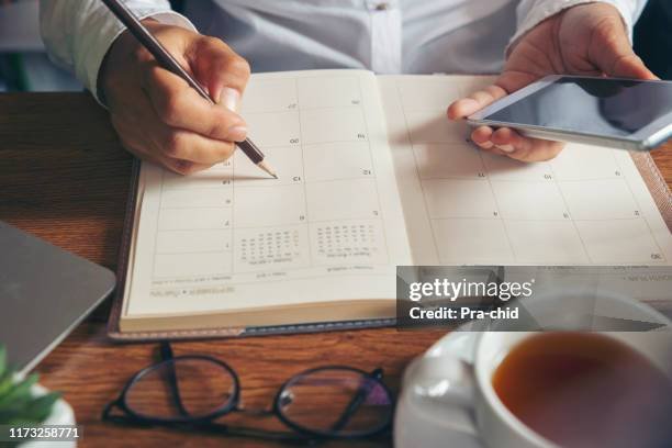 Person writing in a planner with a pen, holding a smartphone, on a wooden table next to a cup of tea or coffee and a pair of glasses.