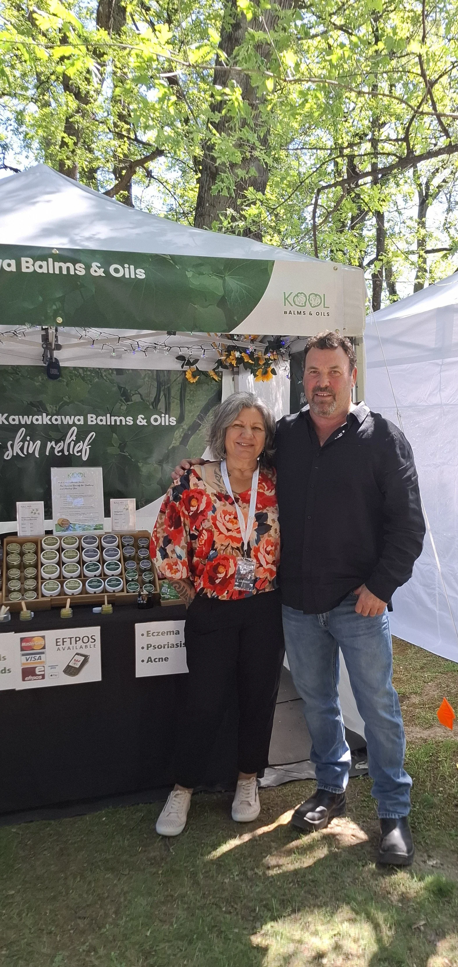 Two people standing in front of a booth in an outdoor setting with green trees. The booth displays wellness products, specifically balms and oils, with signs indicating skin relief, eczema, psoriasis, and acne. The woman on the left wears a red floral top and white sneakers, while the man on the right wears a black shirt and jeans.
