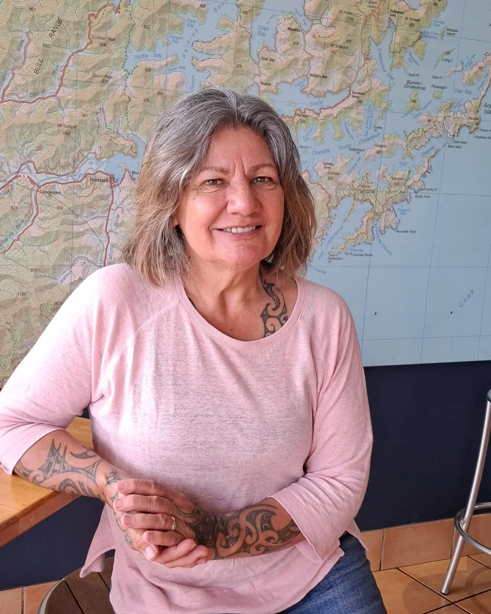 A woman with grey hair smiling, wearing a light pink long-sleeve shirt, with arms crossed, sitting in front of a large map on the wall.