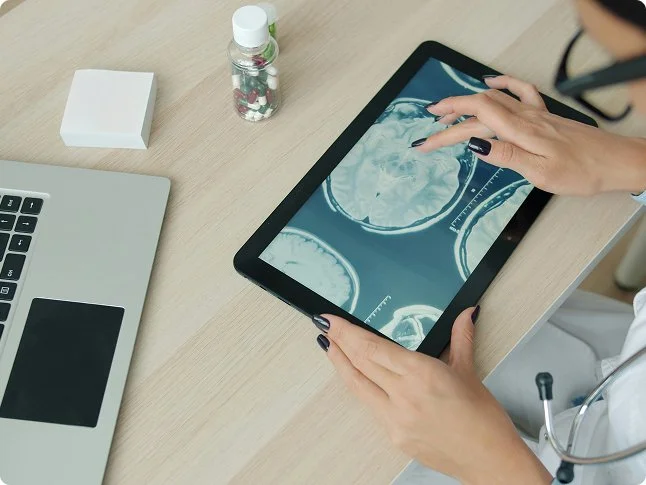 A person in a white coat with a stethoscope examining brain scan images on a tablet. The table also has a laptop, a small bottle of pills, a tissue box, and a pen.