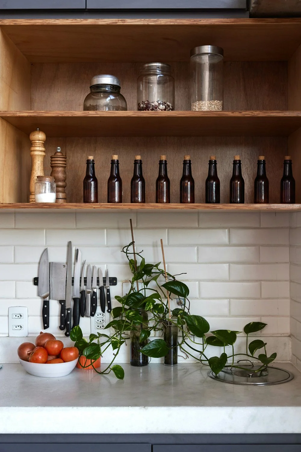 Open kitchen shelf with glass jars and bottles, a white bowl of tomatoes, and a potted plant, on a white brick backsplash.